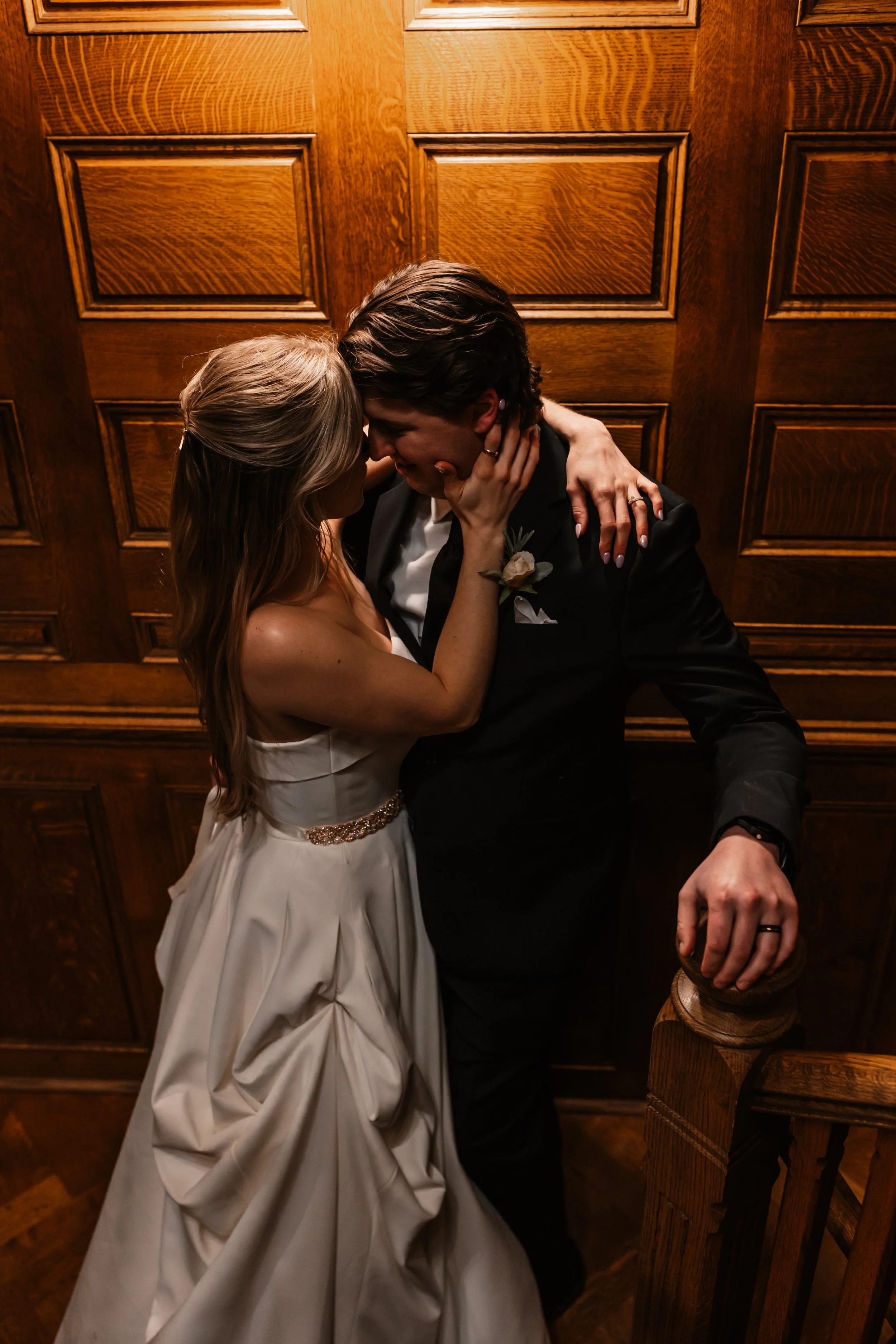 A bride and groom in an embrace, leaning in for a kiss, against a wooden paneled wall, with the groom leaning on a staircase post.