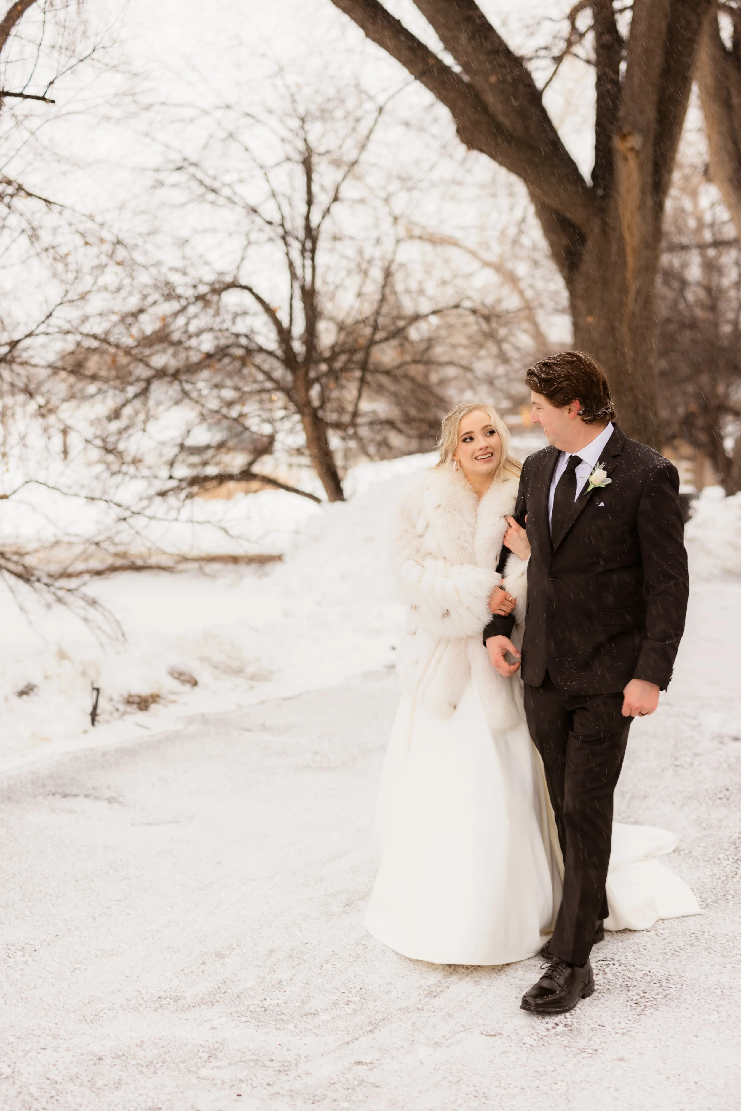 A bride and groom walk arm-in-arm on a snowy path outdoors, surrounded by leafless trees. The bride wears a white wedding gown with a fluffy fur coat, and the groom wears a black suit with a tie and boutonniere. Snow is falling gently around them.