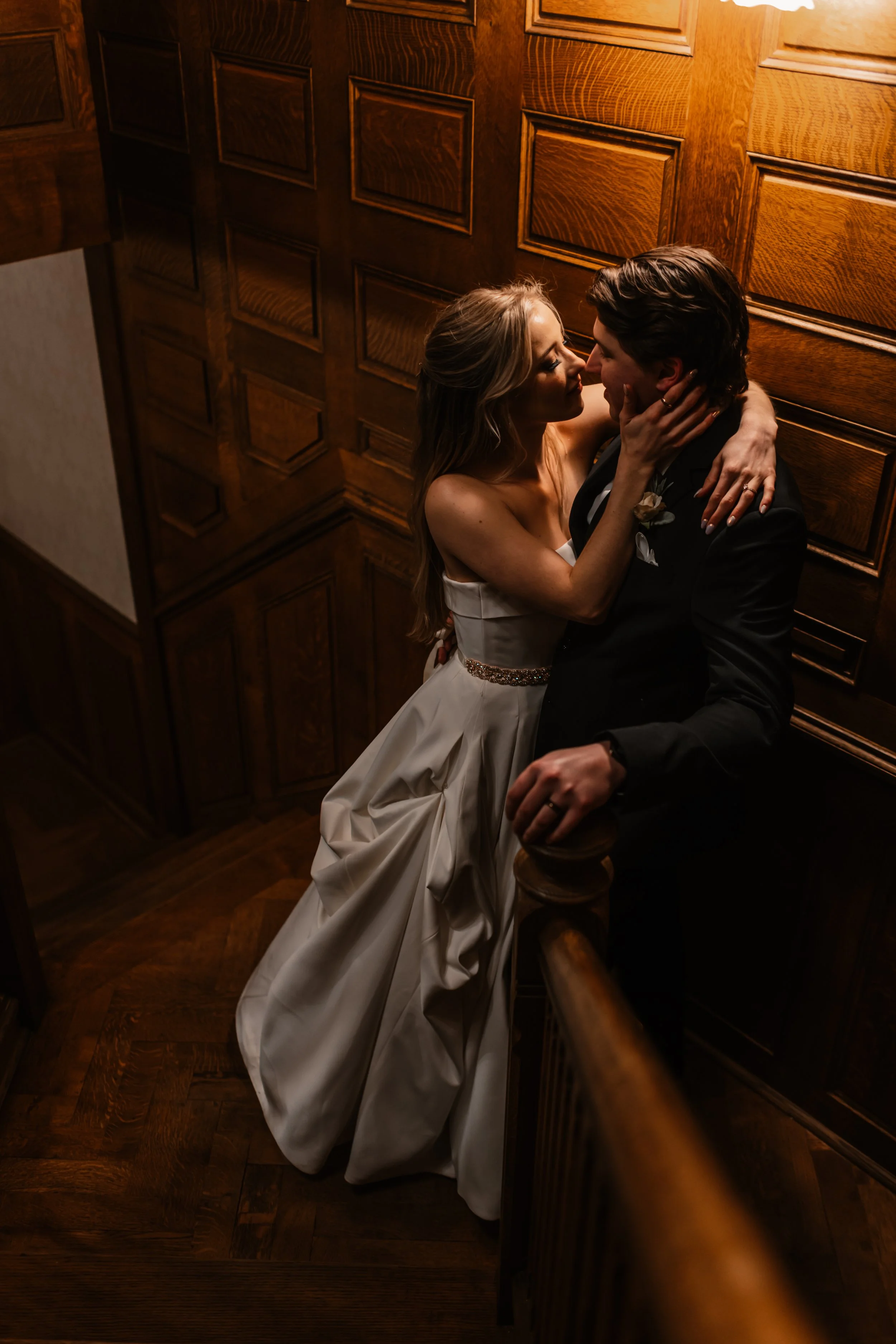 A bride and groom sharing an intimate moment on a staircase with wooden paneling, dressed in wedding attire, with the bride in a white gown and the groom in a black suit.