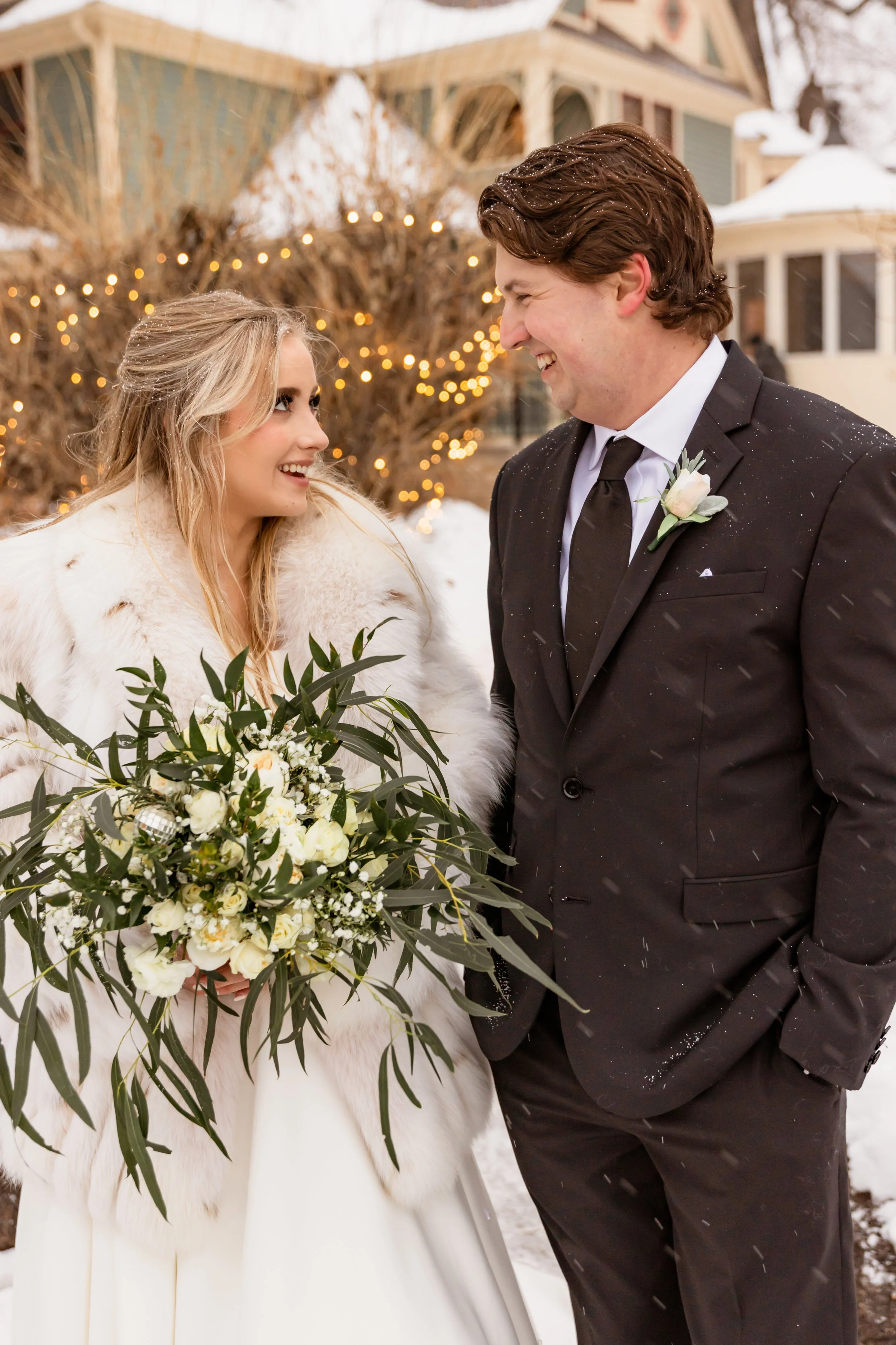 A bride and groom standing outside in the snow, smiling at each other during their winter wedding, with string lights and a house in the background.
