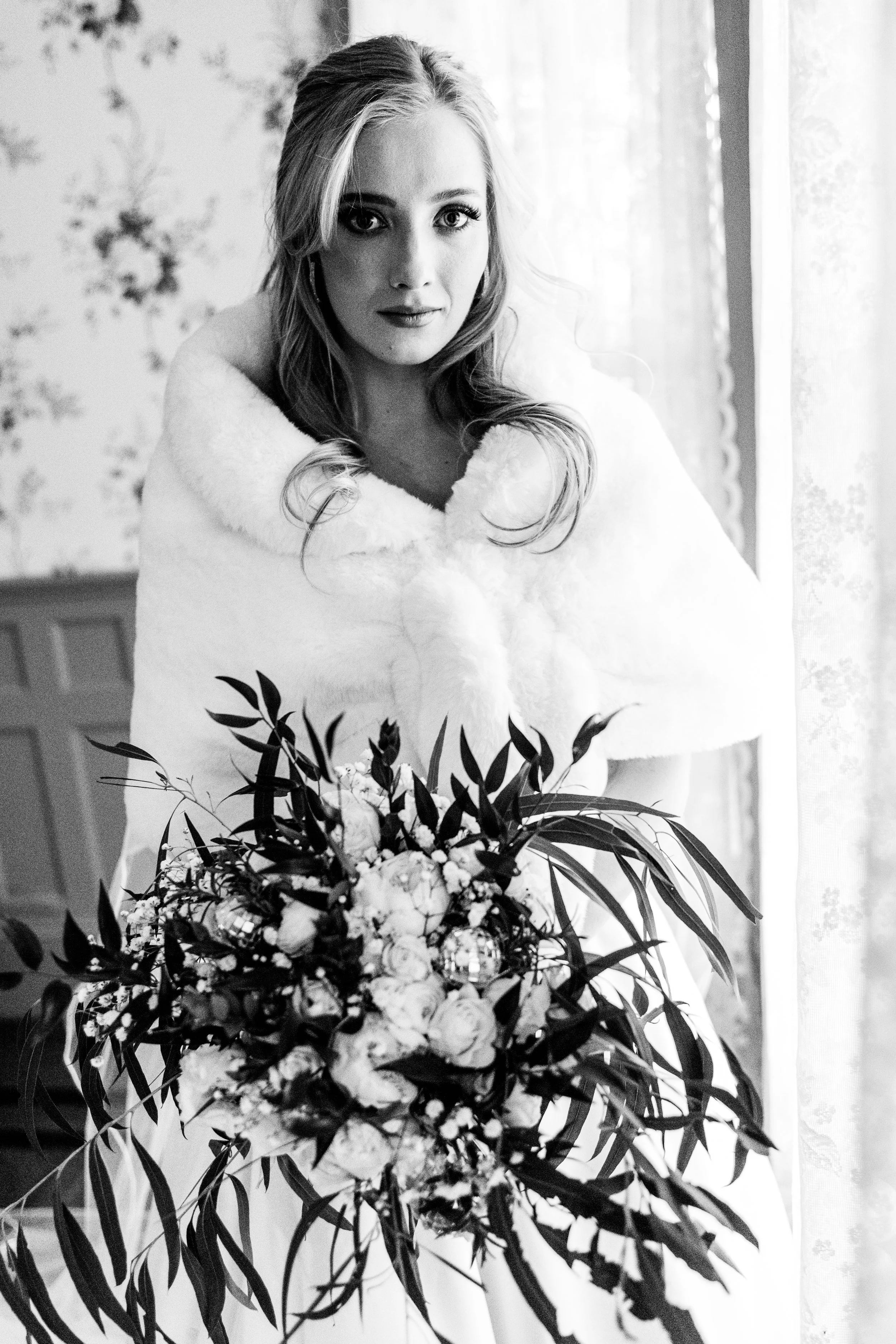 Black and white photo of a young woman with blonde hair and expressive eyes, wearing a white fur shawl, holding a large bouquet of flowers, standing near a window with lace curtains.
