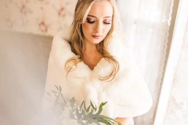 A woman with blonde hair wearing a white, fluffy coat sitting by a window with sheer curtains, holding a bouquet of green leaves.