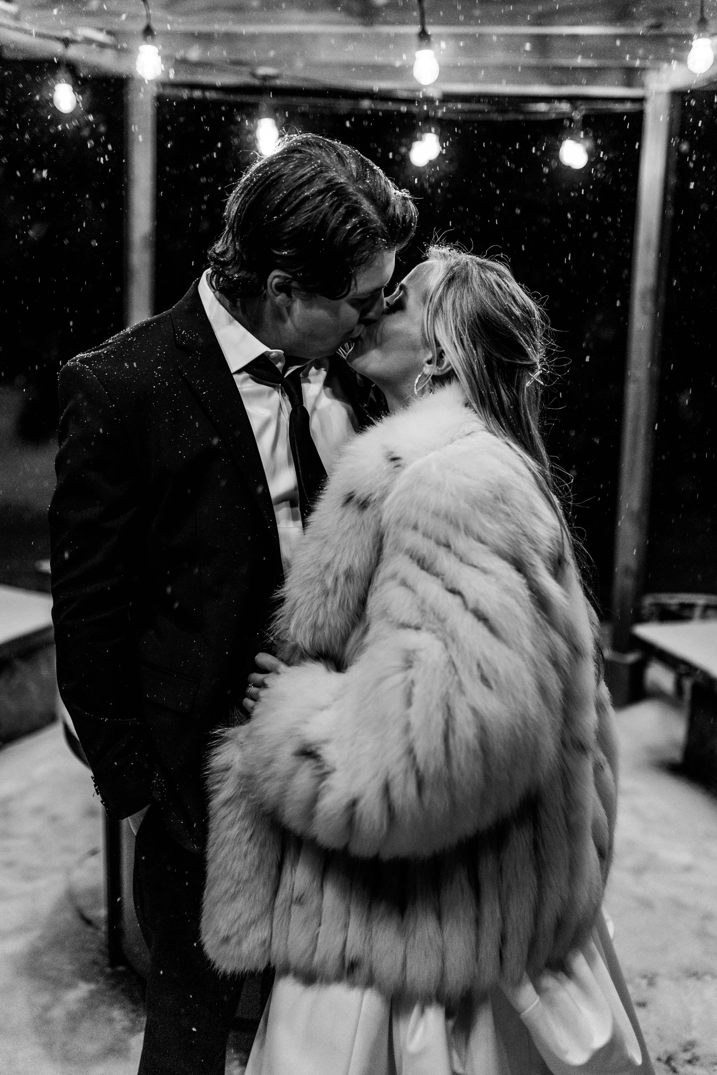 A black and white photo of a couple kissing outdoors at night, with string lights overhead and snow falling.