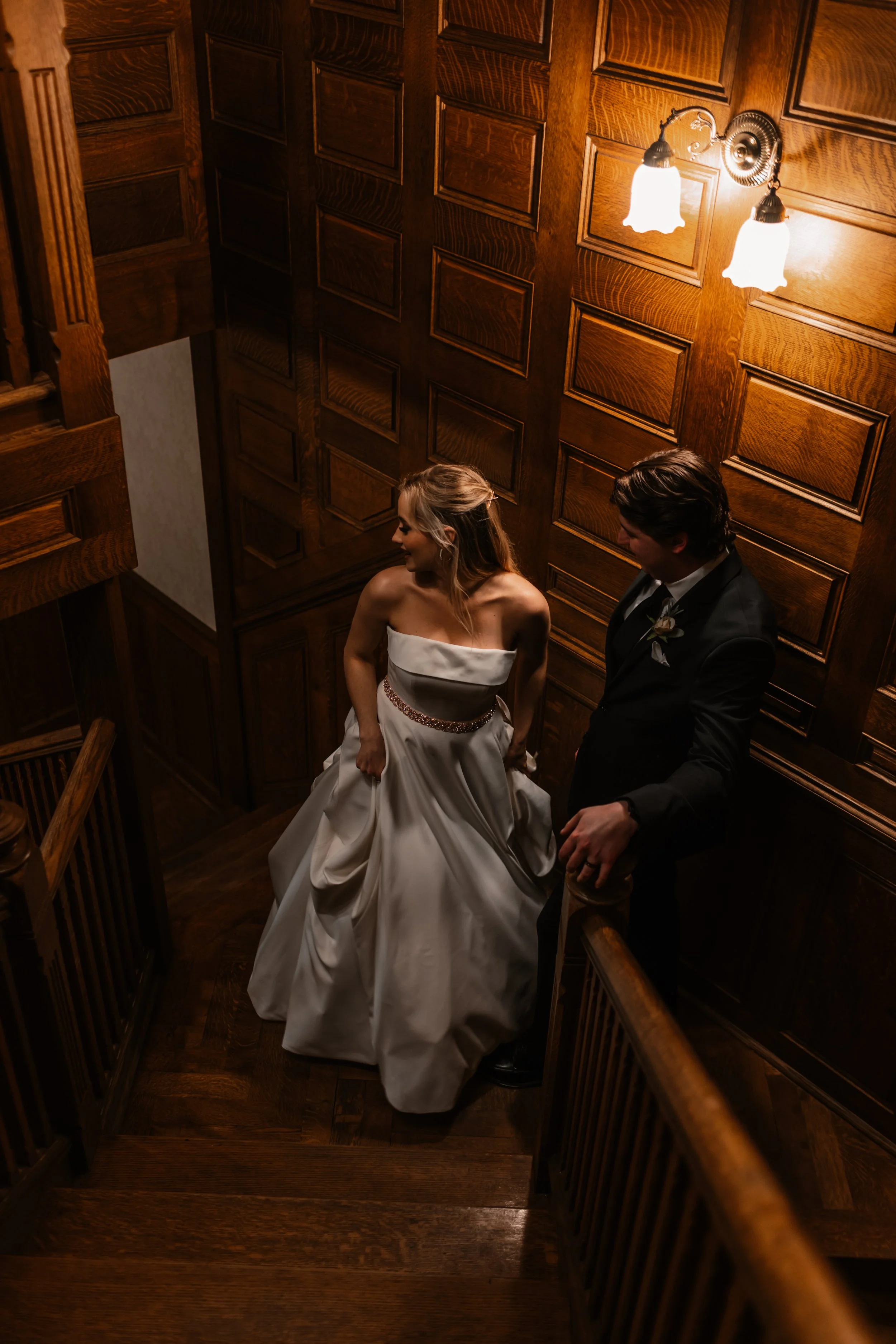 A bride and groom standing at the top of a wooden staircase in a dimly lit, wood-paneled hallway, sharing a moment. The bride is wearing a strapless white wedding gown, and the groom is in a black suit. Warm lighting from two wall sconces illuminates