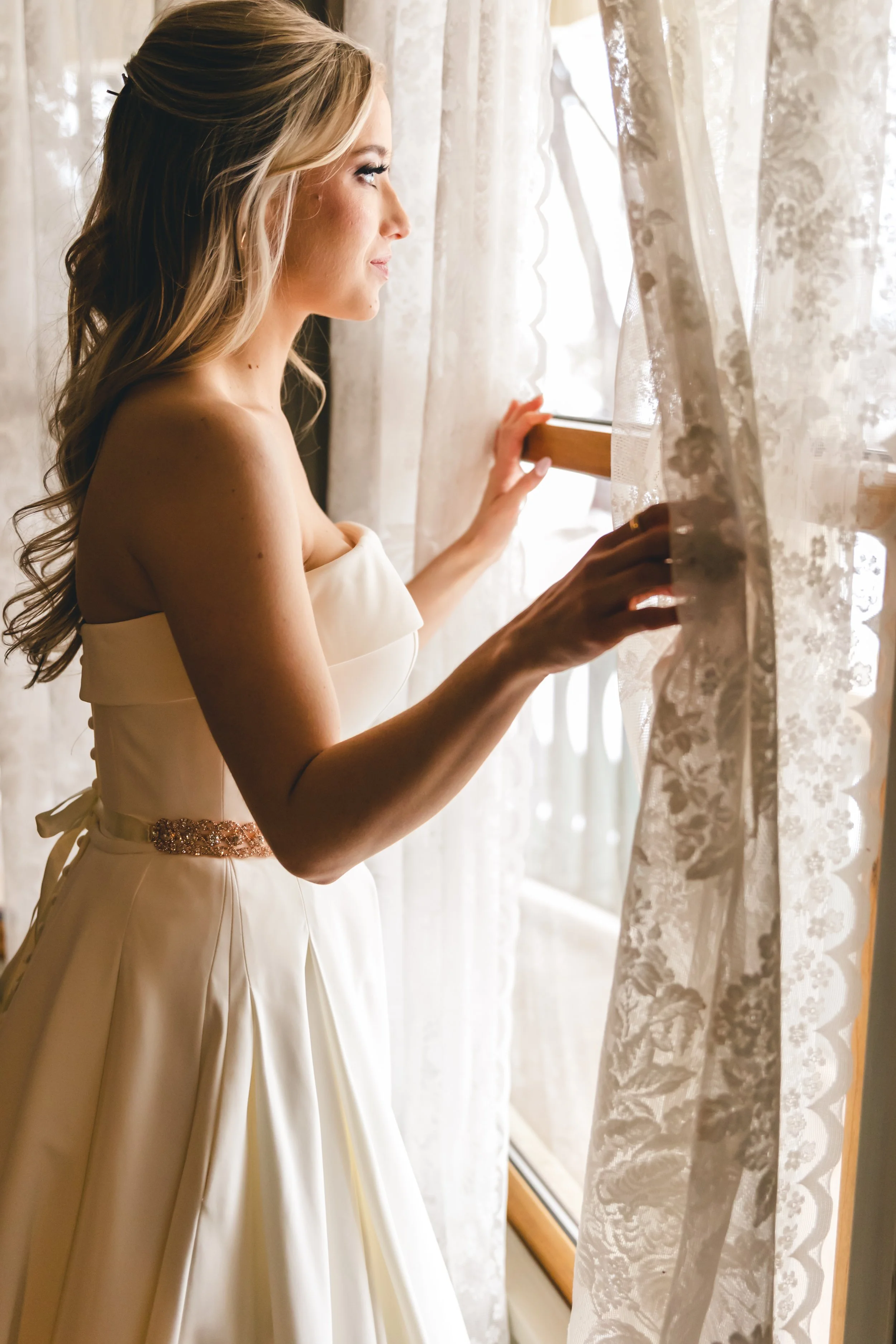 A bride in a white wedding dress looks out a window through lace curtains.