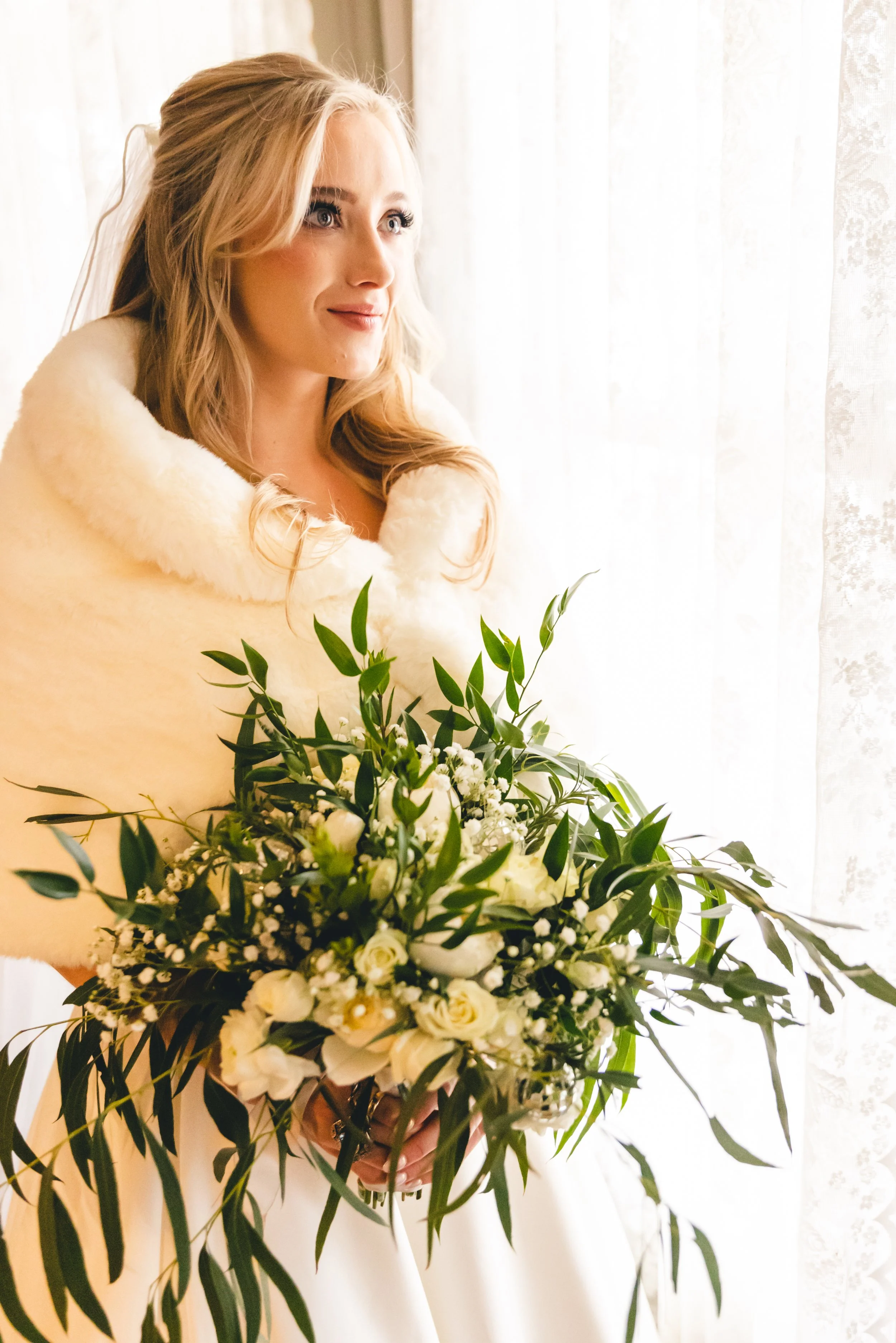 Bride with blonde hair in a white dress and fur shawl holding a large bouquet of white roses and green leaves, looking out a window with sheer curtains.