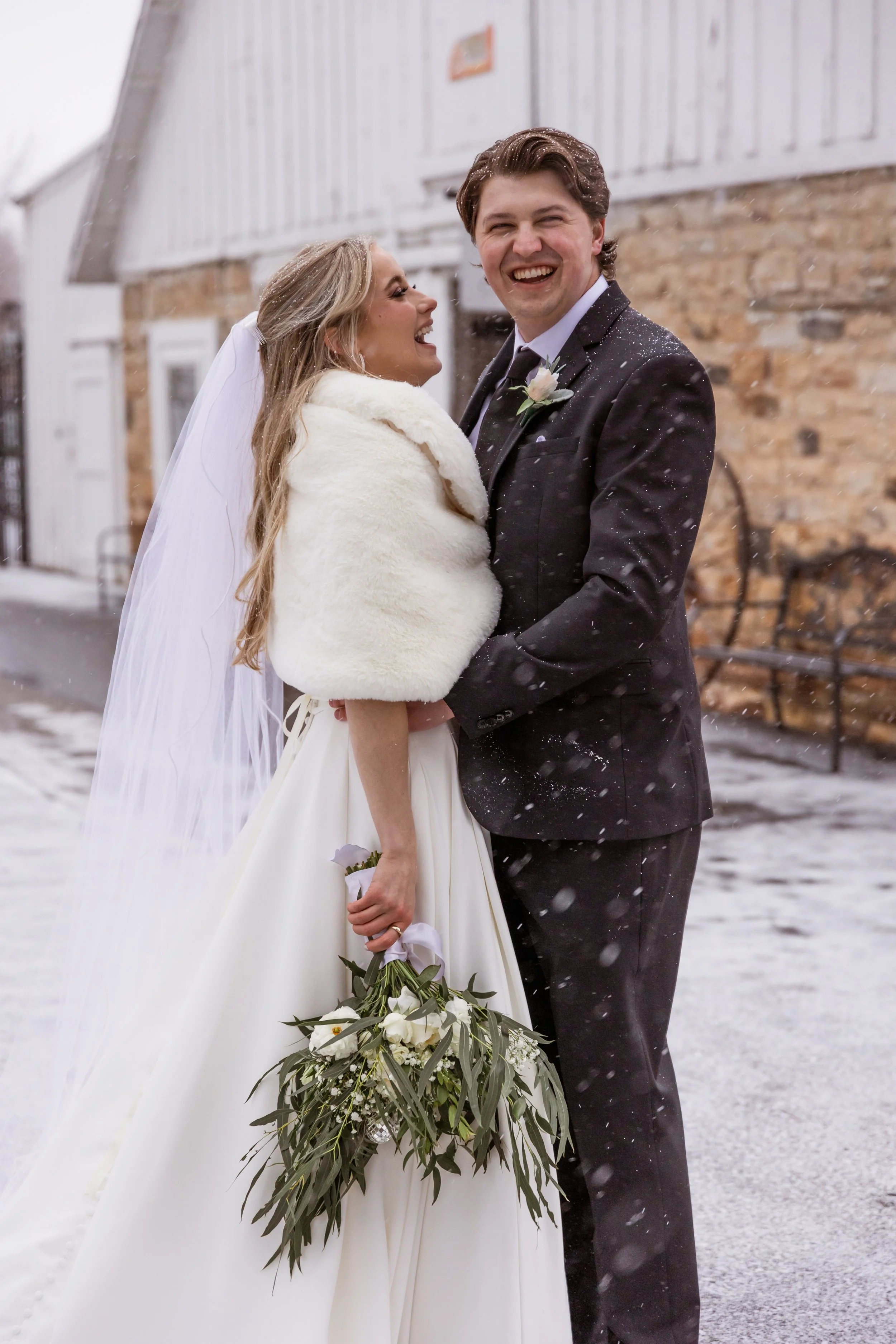 A bride and groom laughing outside on a snowy day, with a rustic brick and wood building in the background. The bride is wearing a white gown, a fur stole, and holding a bouquet of white flowers, while the groom is in a black tuxedo with a boutonnier