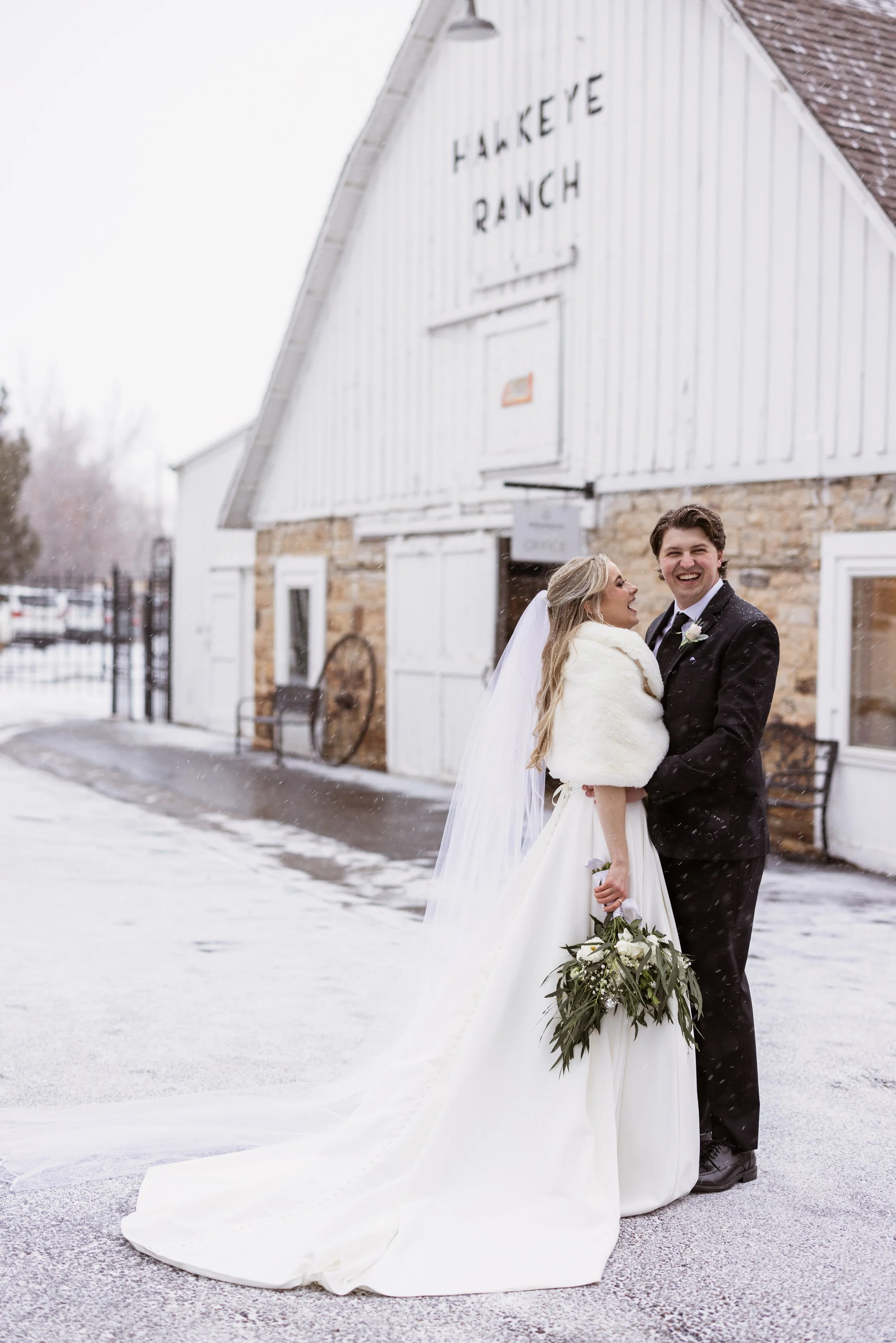 A bride and groom smiling and holding hands outside in the snow, with a rustic barn labeled 'Hakkeve Ranch' in the background.