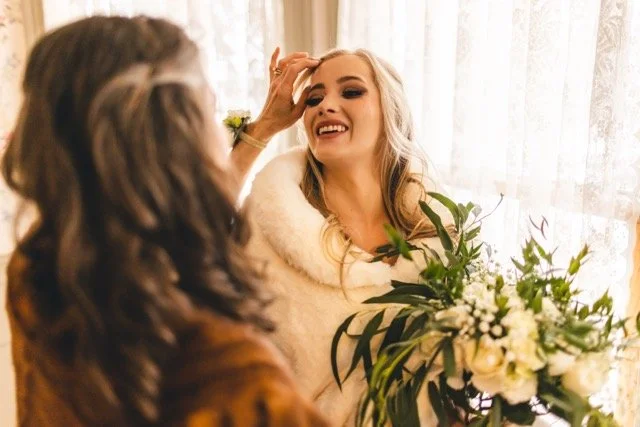 A woman with long blonde hair smiling and holding a bouquet of flowers, while another woman with dark hair, back to camera, touches her forehead in a bright room with curtains.