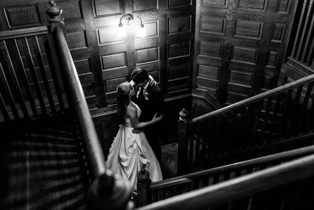 A couple in wedding attire sharing a kiss in a dimly lit, wood-paneled stairwell.