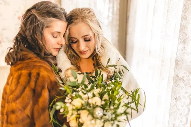Two women, one with dark hair and one blonde, holding a bouquet of flowers, standing close together by a window with lace curtains.