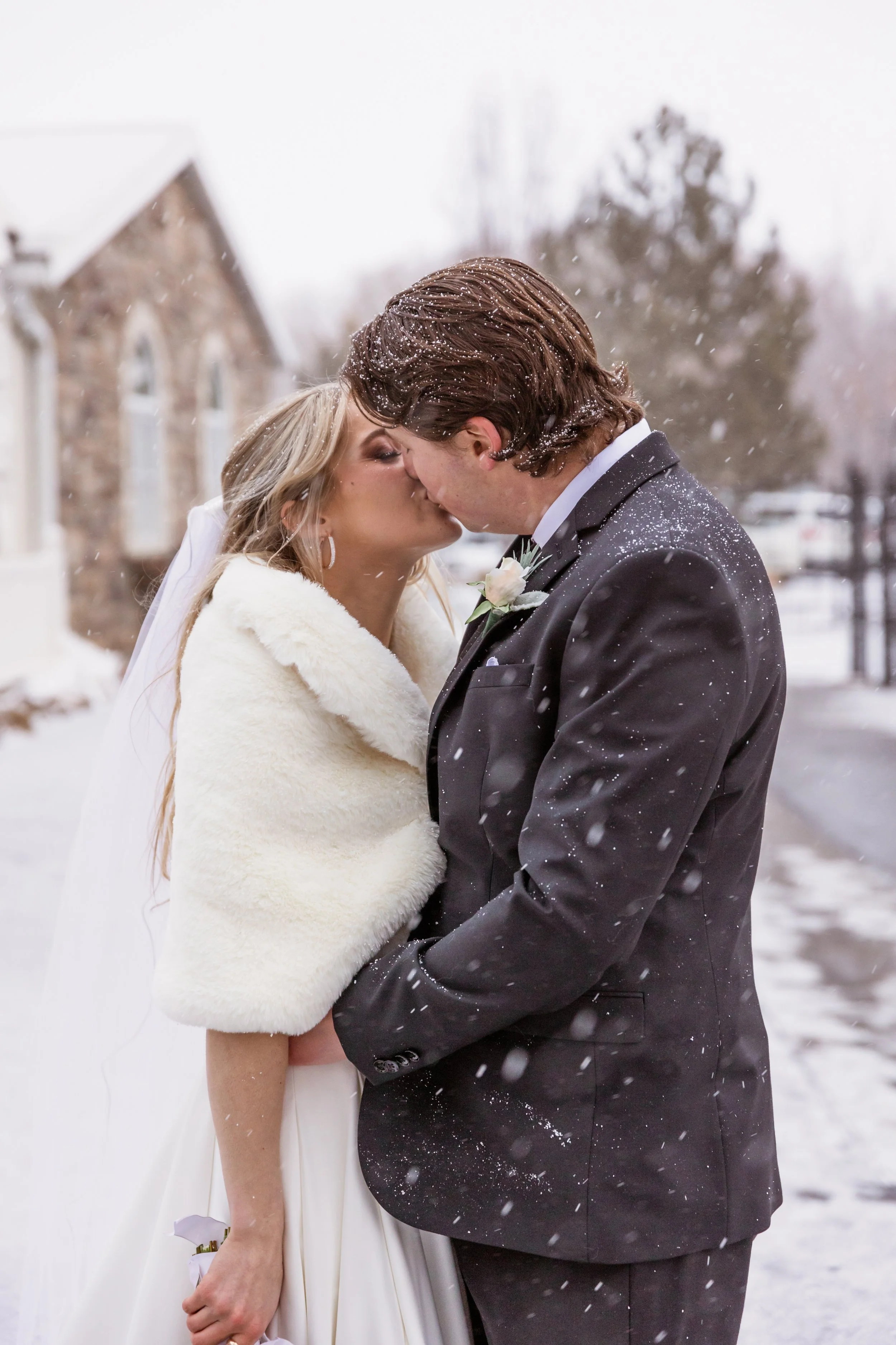 A bride and groom share a kiss outdoors in snowy weather during their wedding day.
