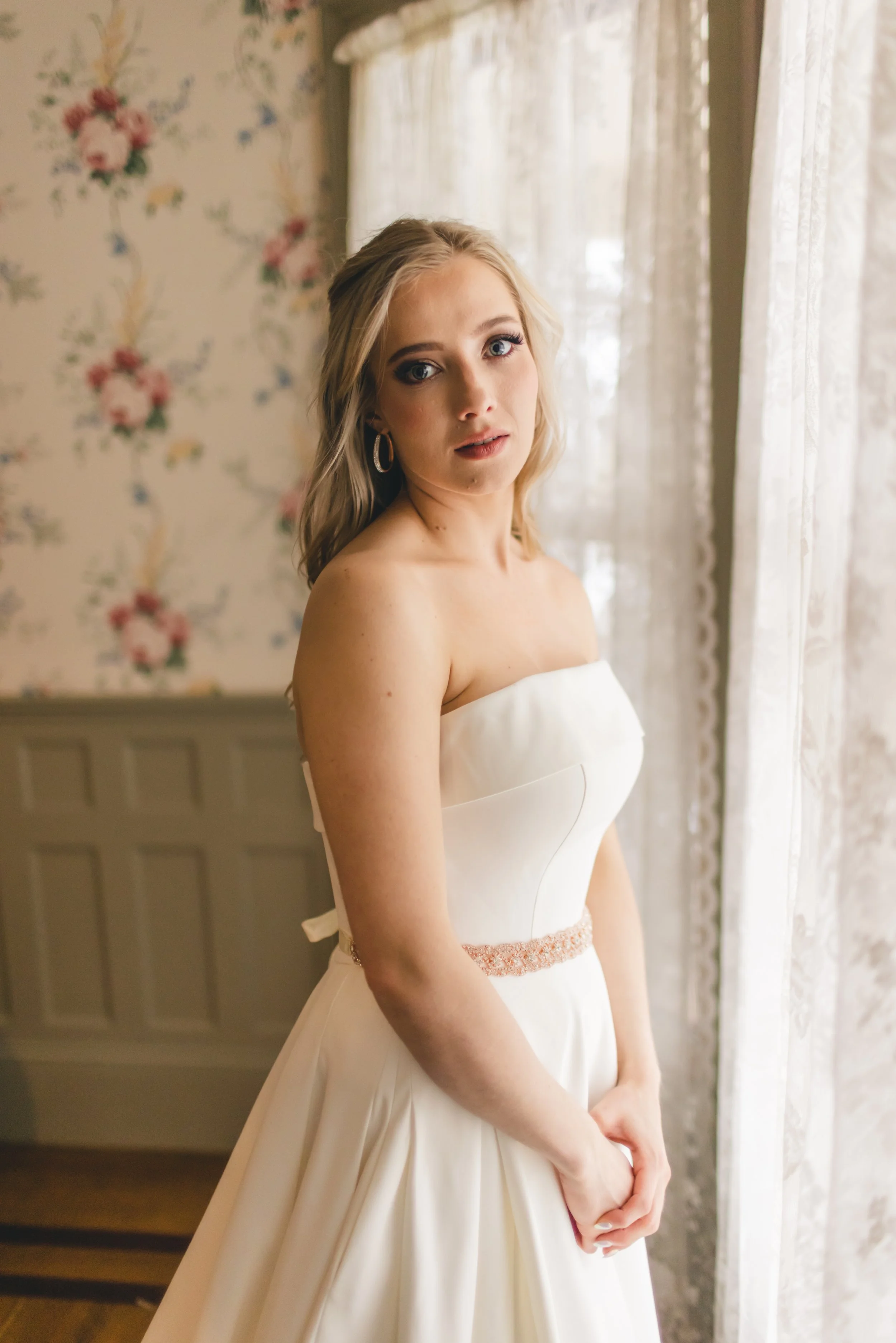 A young woman in a strapless white wedding dress standing near a window with lace curtains, looking at the camera with a neutral expression.