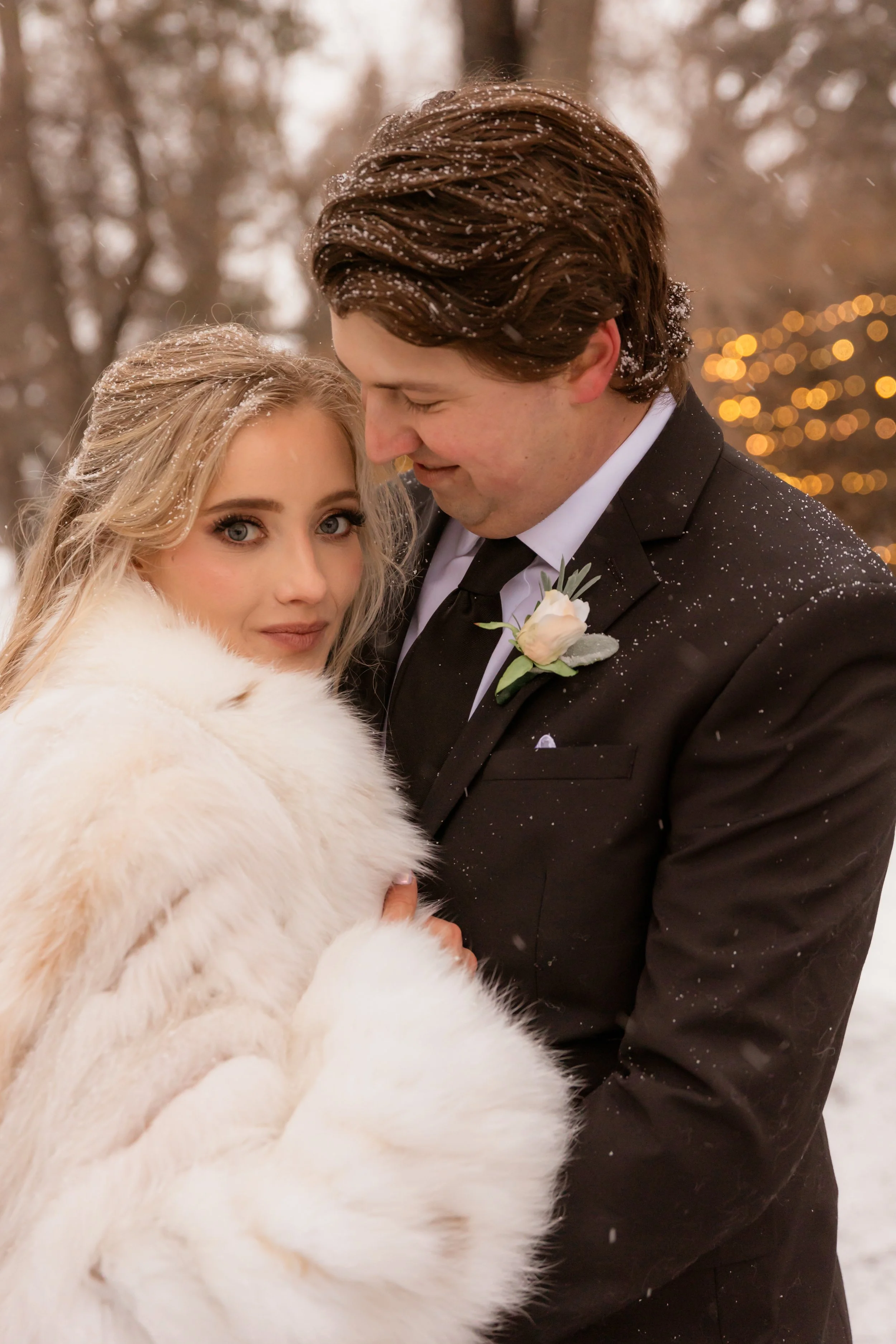 A bride and groom standing close together outdoors on a snowy day, with the bride wearing a white fur coat and the groom in a black suit with a white boutonniere, both smiling softly.