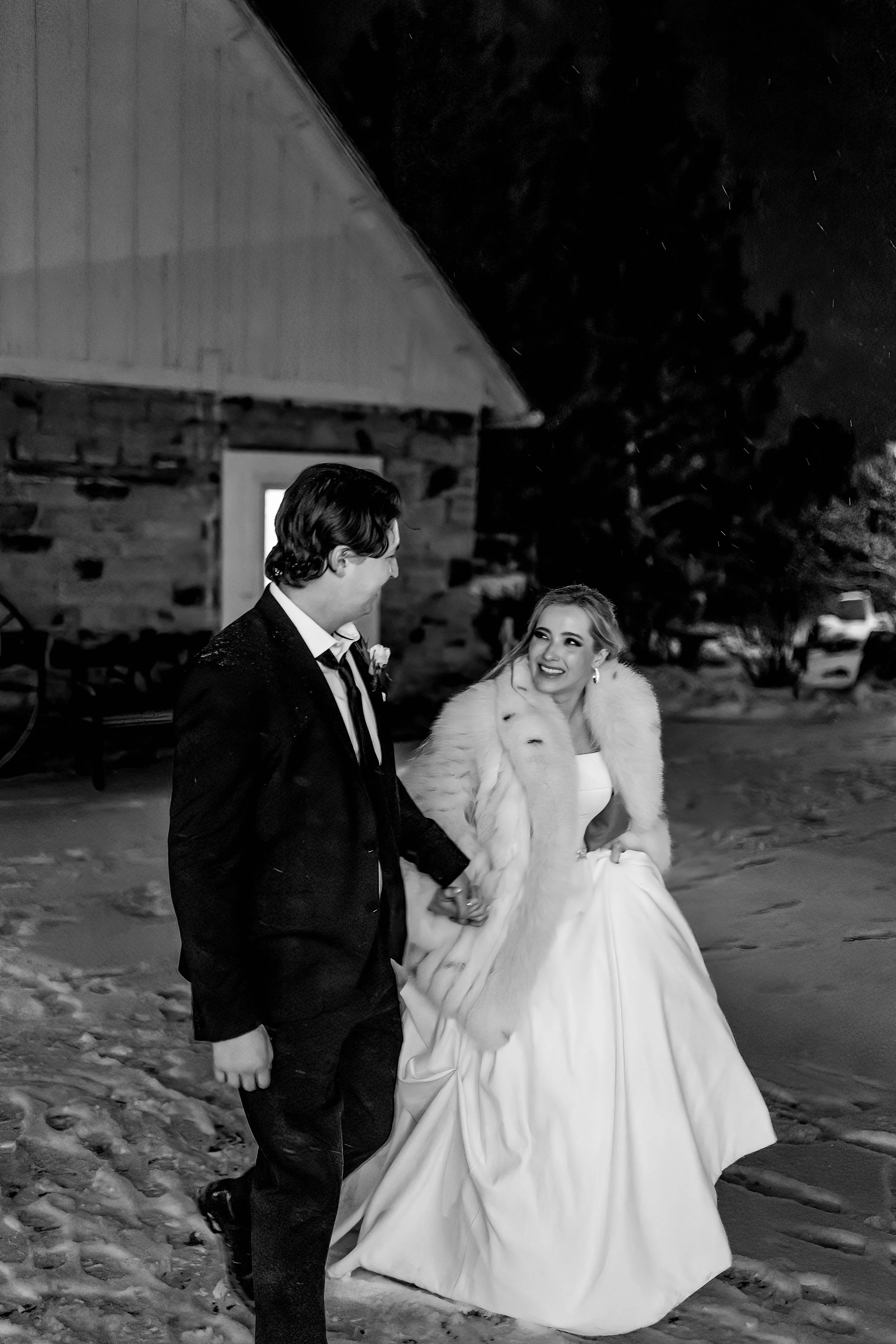 A couple dressed in wedding attire, holding hands and smiling at each other outdoors at night, in a snowy setting with a rustic building in the background.