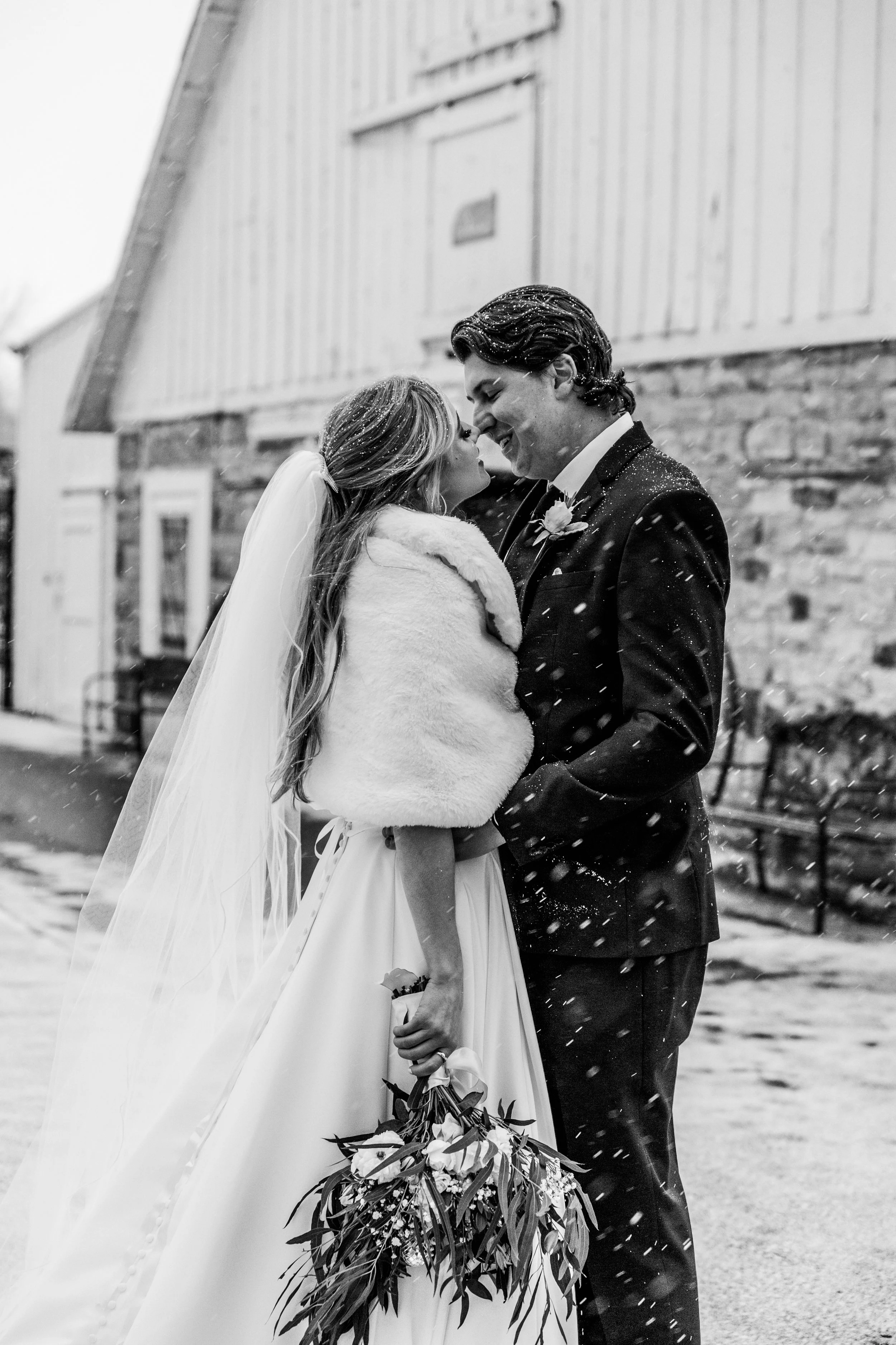 A black and white photo of a bride and groom smiling and holding hands on their wedding day, outdoors in the snow, with a rustic building in the background.