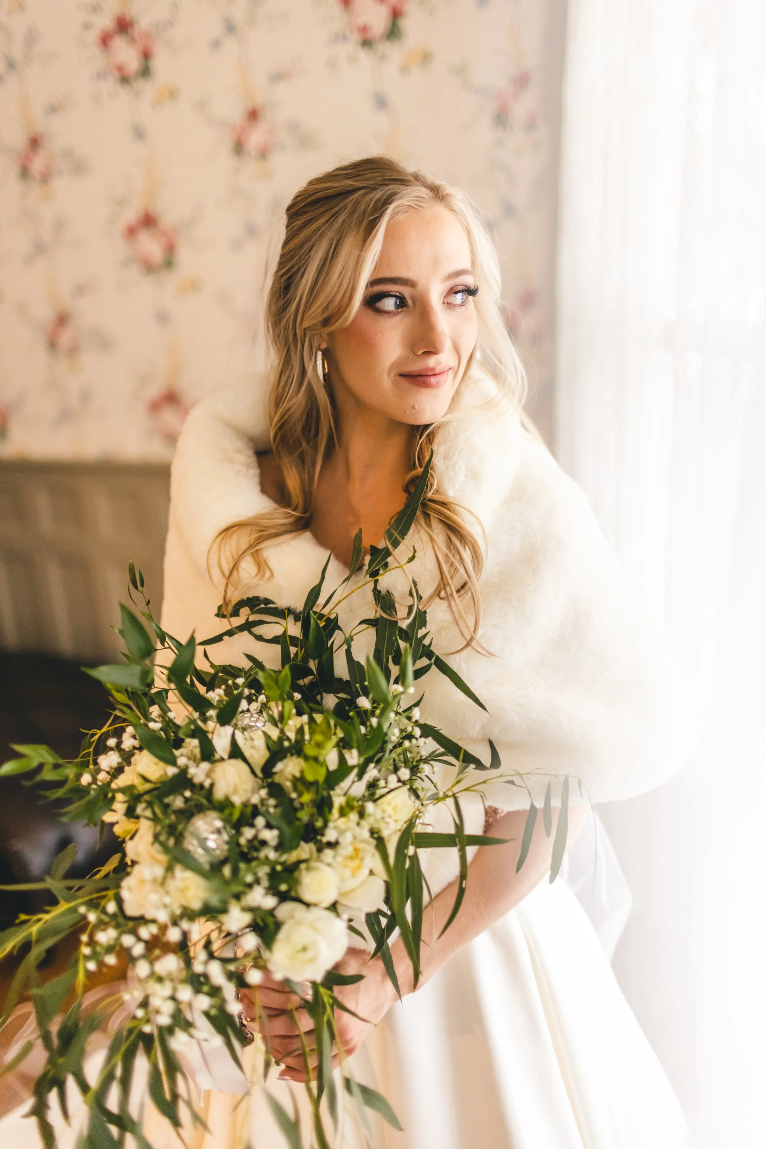 A bride in a wedding dress holding a bouquet of white flowers and greenery, standing by a window with floral wallpaper in the background.