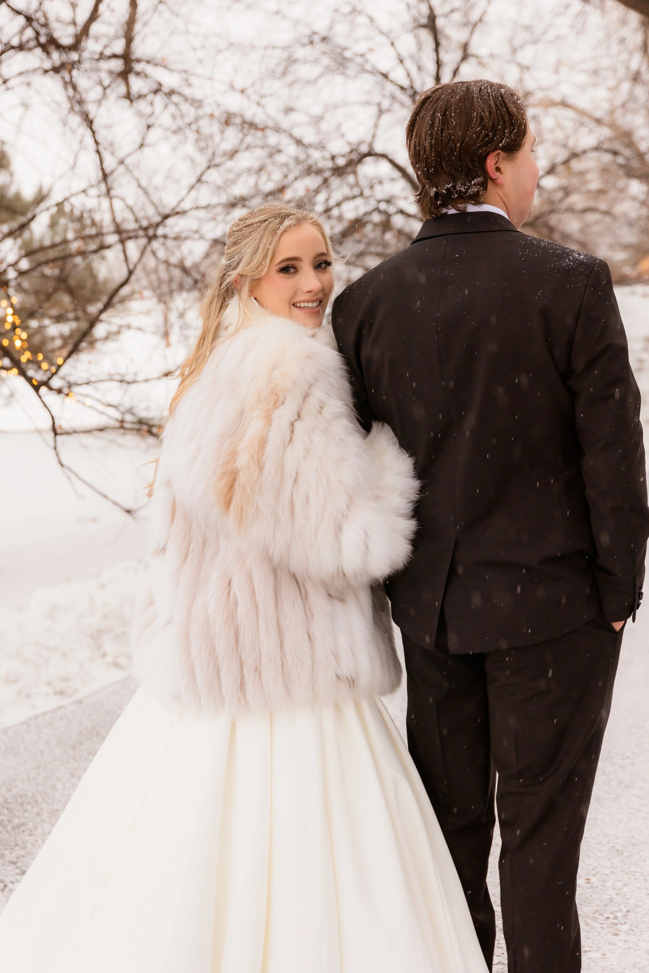 A smiling woman in a wedding dress and fur coat stands next to a man in a black suit outside in a snowy landscape, with leafless trees and holiday lights in the background.