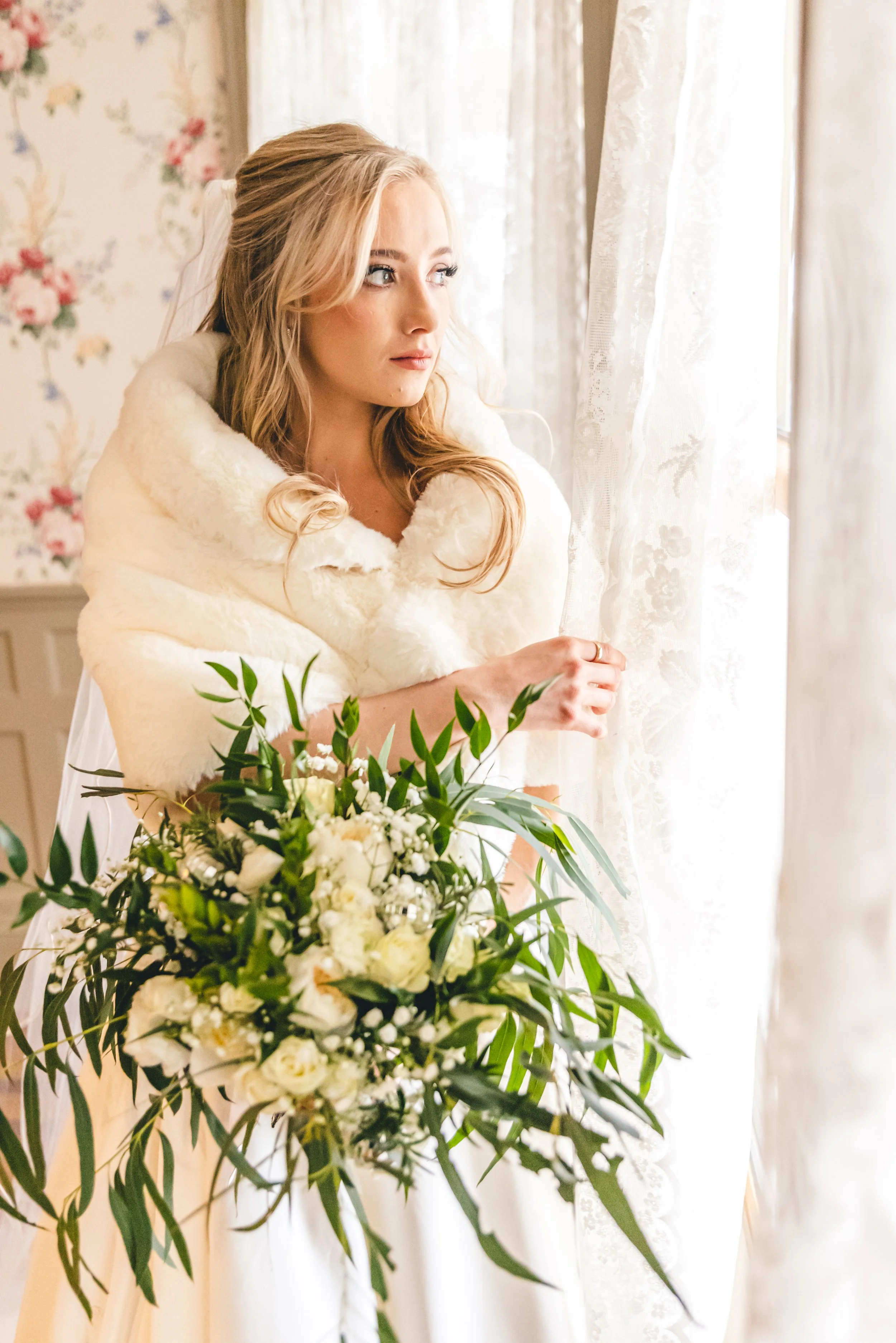 Bride looking out a window, holding a bouquet of white flowers and greenery, wearing a faux fur wrap.