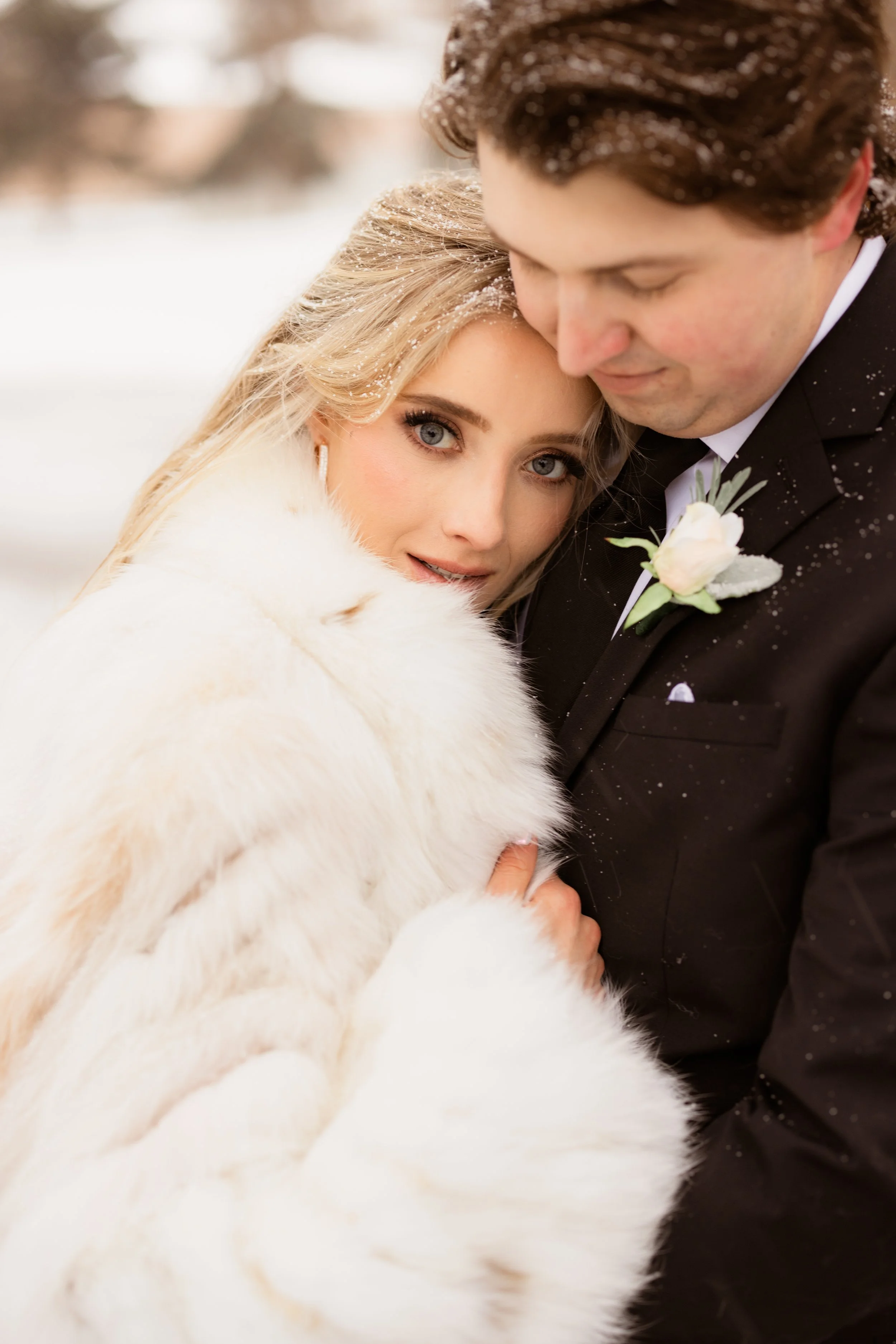 A bride and groom close together on a snowy day, with the bride wearing a white fur coat and the groom in a black tuxedo. The bride has blonde hair and blue eyes, looking at the camera, while the groom leans his head down, with snowflakes on their ha
