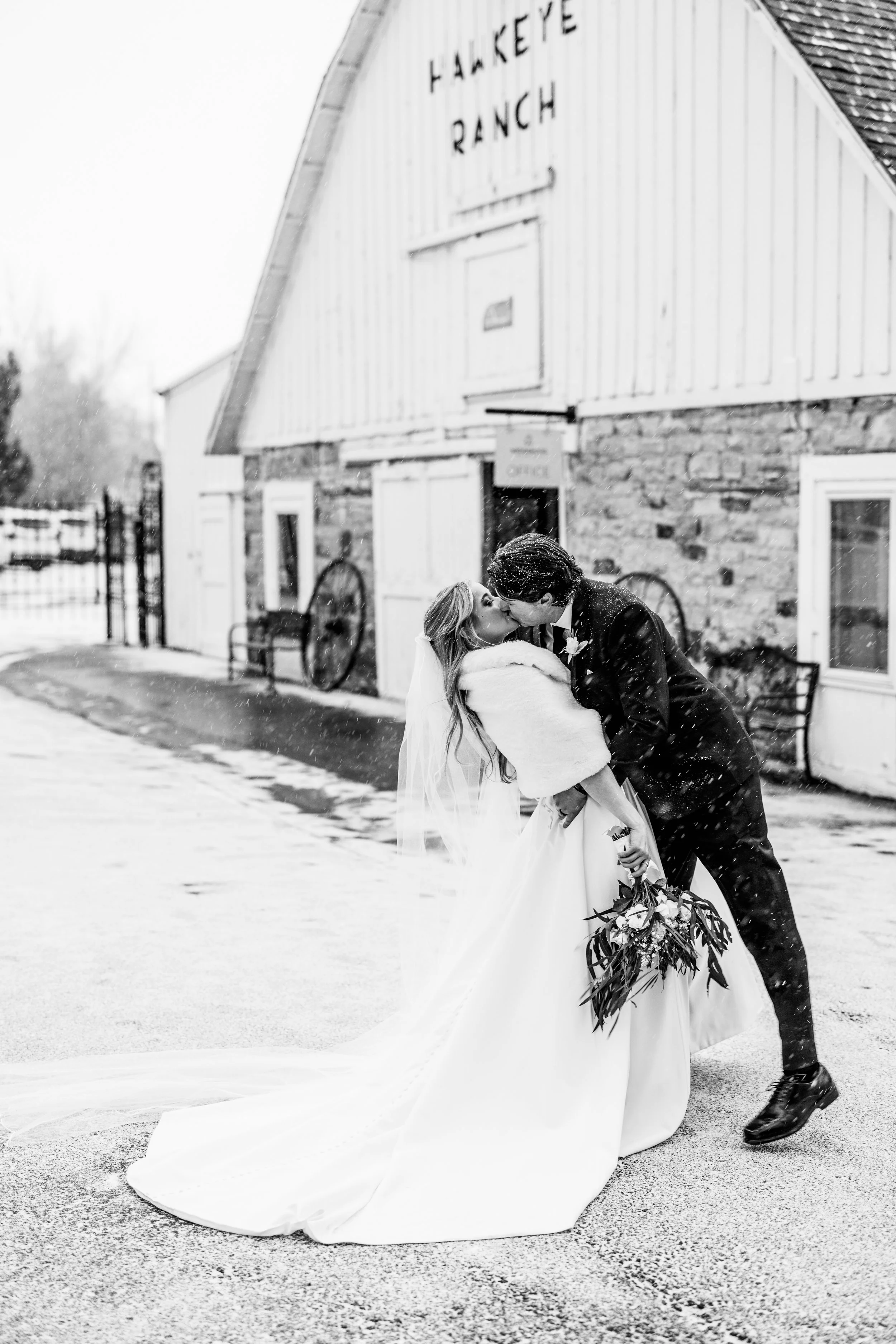 A newlywed couple sharing a kiss outside Hancock Ranch, with snow falling around them, in black and white.