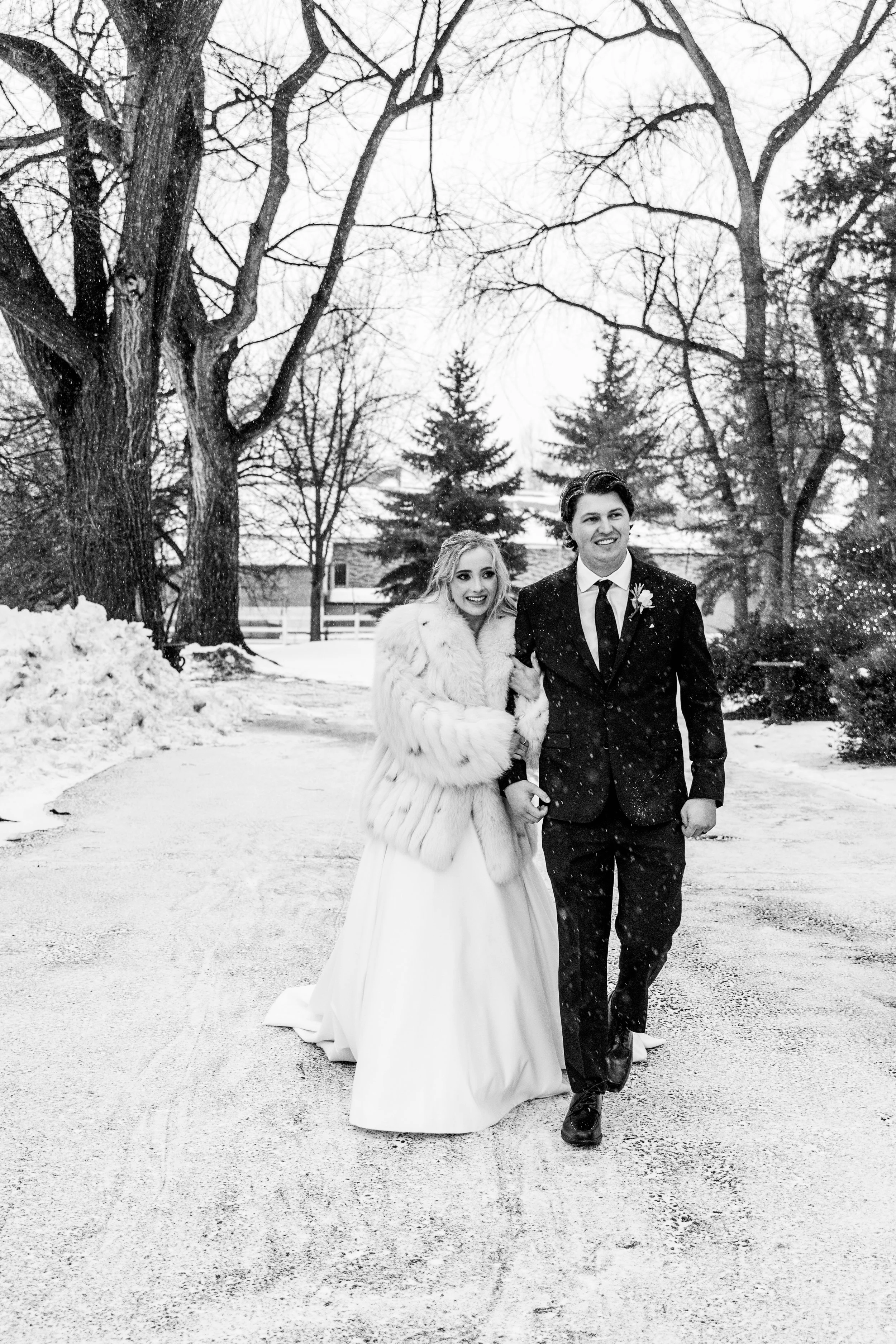 Black and white photo of a smiling bride and groom walking outdoors in a snowy landscape, surrounded by bare trees and evergreens.