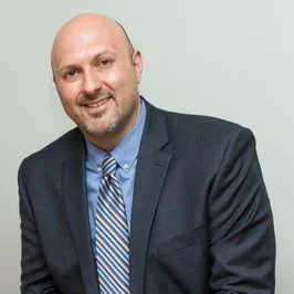 Headshot of a man with a beard, wearing a dark suit and a blue shirt with a striped tie, smiling at the camera.