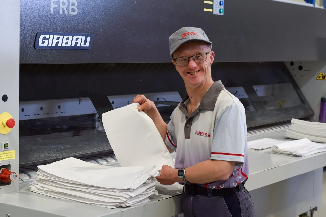 Supported employee folding a white pillow case. He is smiling at the camera and standing in front of a laundry machine.