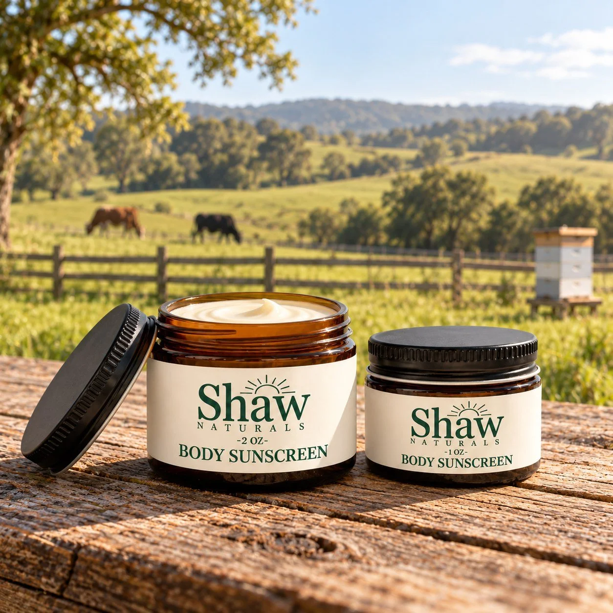 Open and closed Shaw Naturals sunscreen jars on a farm table with pasture, cows and a beehive beyond