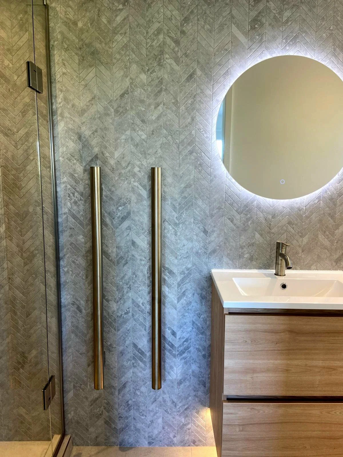 Modern bathroom with a round LED mirror, gray textured tile wall, wooden vanity with a white sink, and brass vertical handles.