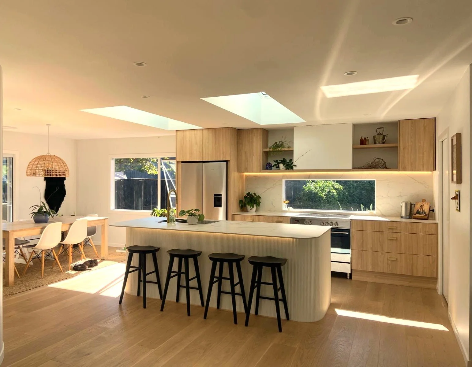 Modern kitchen with light wood cabinets, white countertops, and a central island with black bar stools. Skylights illuminate the room, and a dining table with white chairs is visible on the left. Potted plants and kitchen appliances are neatly arranged.