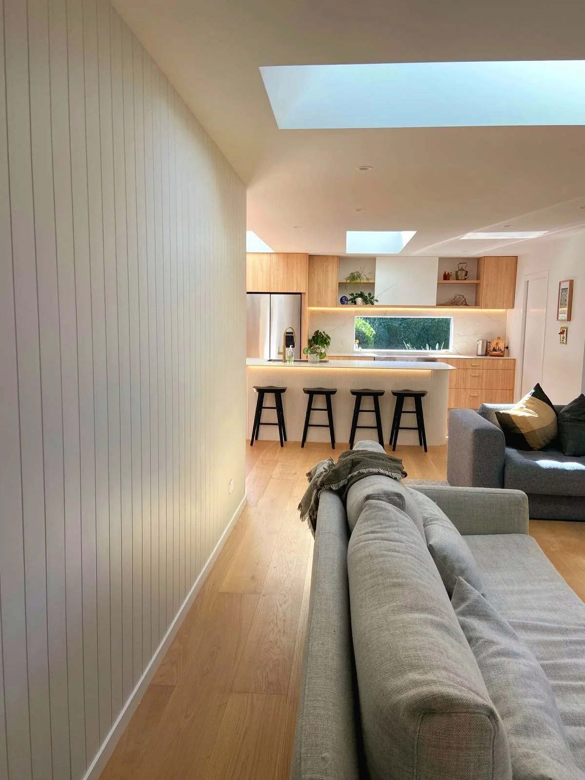 Modern open plan living room with gray sofa, light wooden flooring, a kitchen island with black bar stools, skylights, and wooden kitchen cabinets.