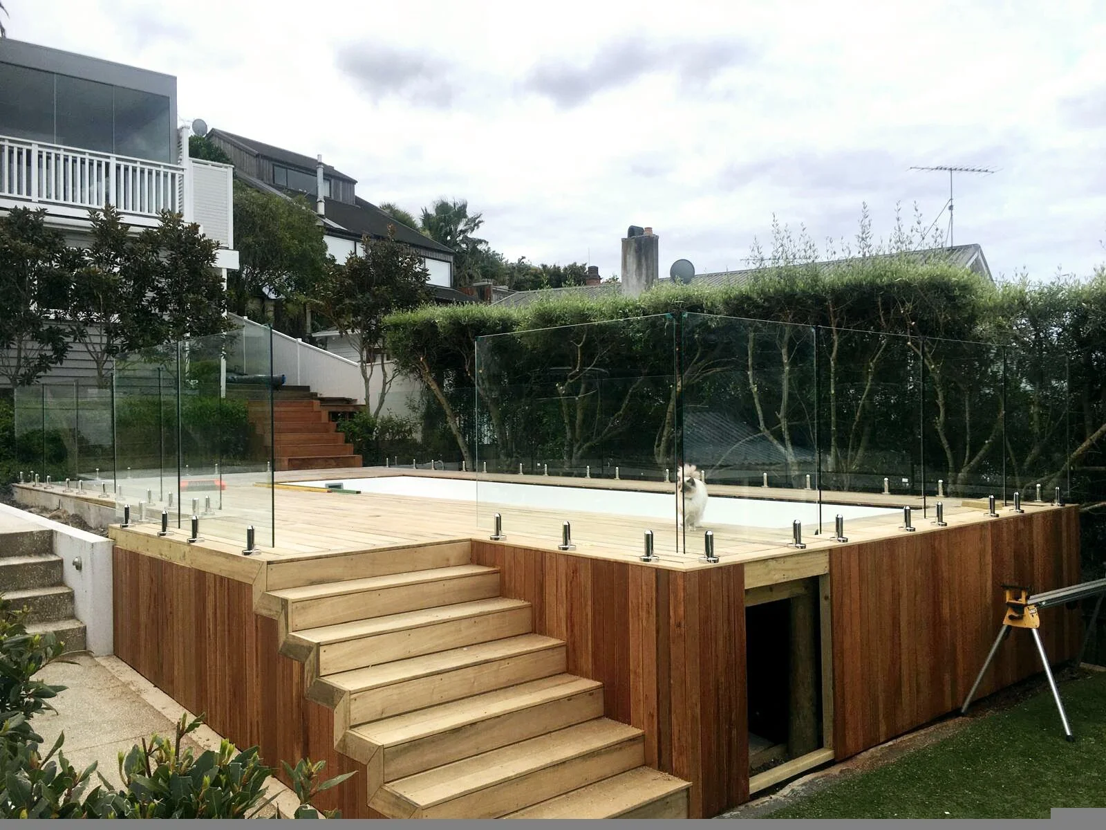 Wooden deck with glass railing around an empty pool, surrounded by greenery and residential buildings in the background.