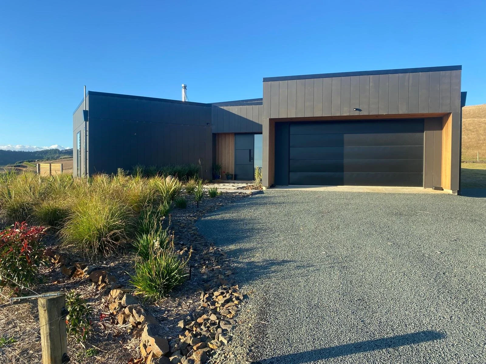 Modern single-story house with dark exterior, large garage door, gravel driveway, and landscaped garden under clear blue sky.