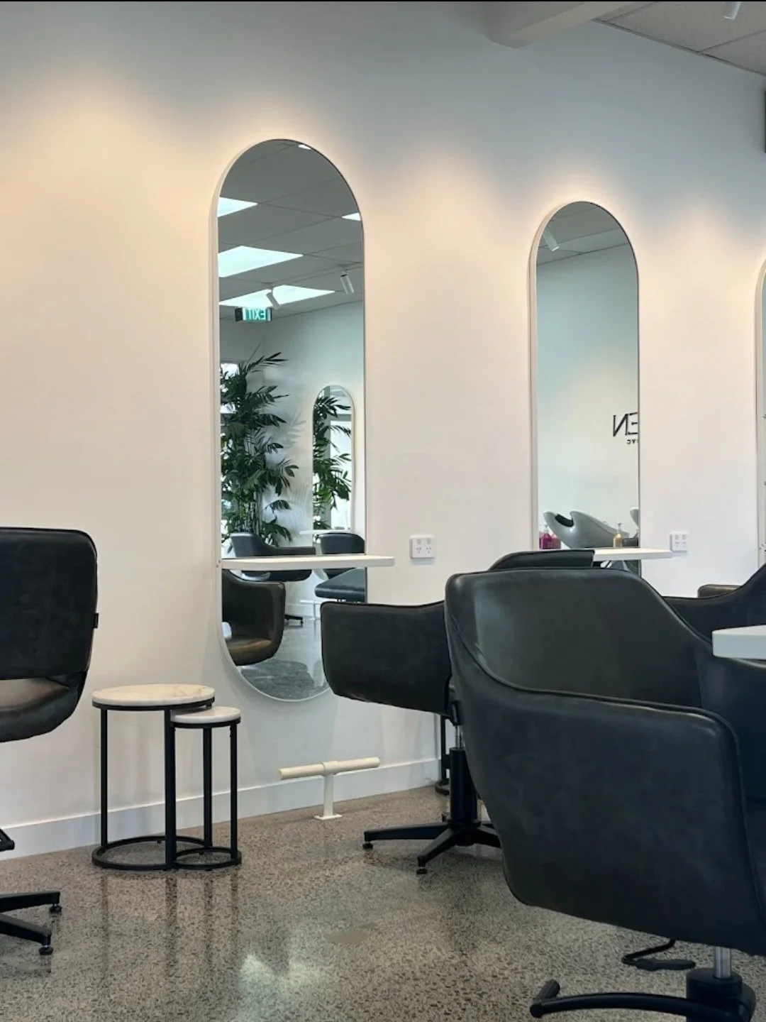 Modern hair salon interior with black chairs, round mirrors, and marble-top tables. The room features large arch mirrors and potted plants, creating an upscale and stylish atmosphere.