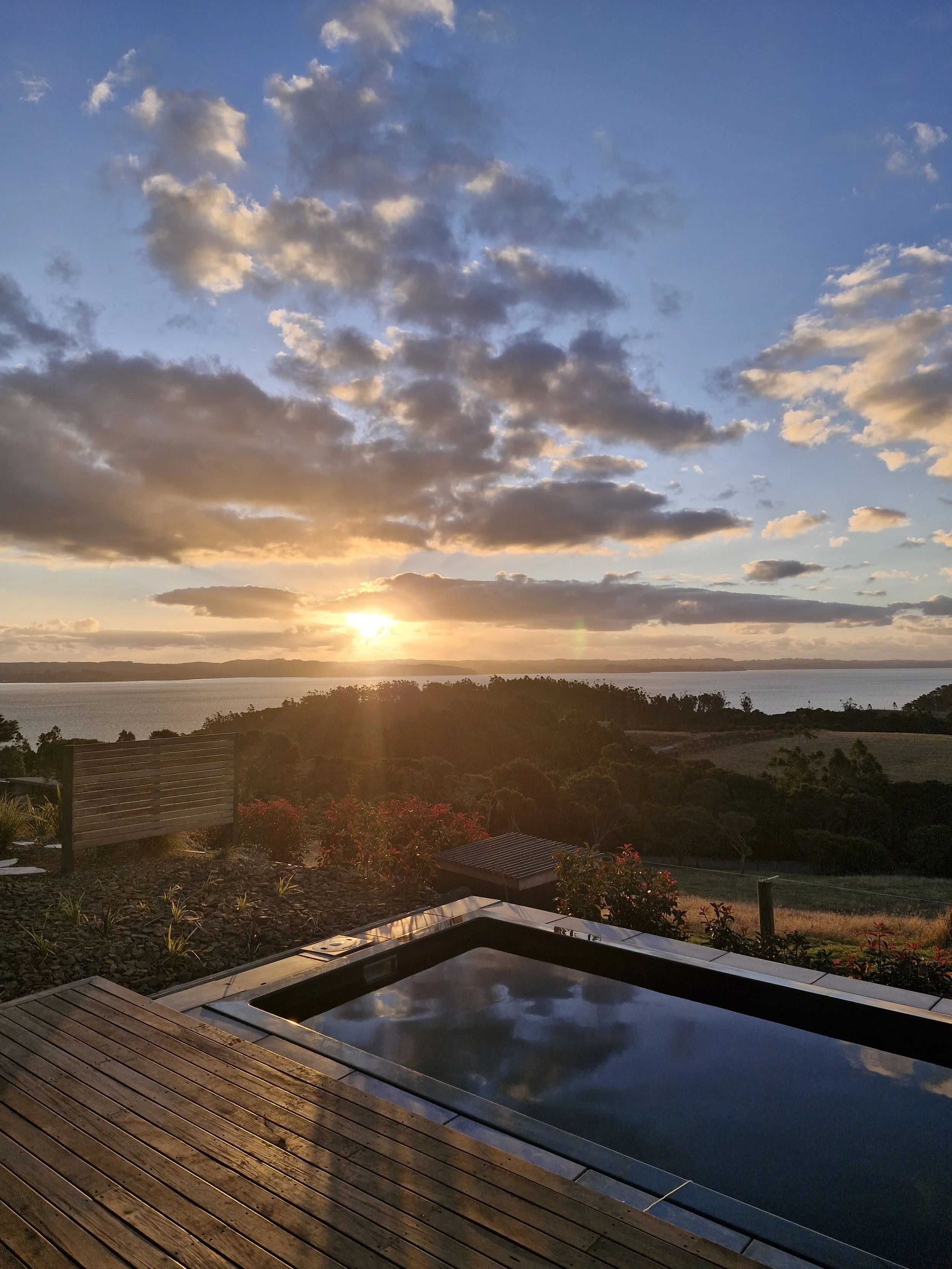 Scenic sunset view over a calm pool with wooden decking, surrounded by lush greenery and red foliage, overlooking a distant body of water and hills under a cloudy sky.