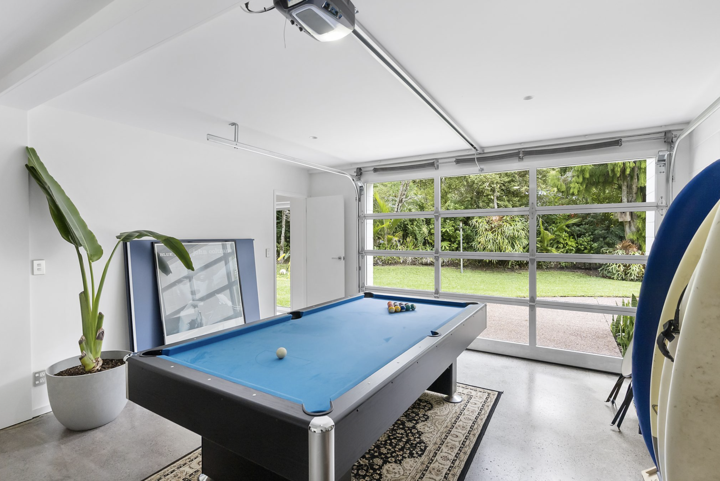 A well-lit garage with a blue pool table, surfboards stacked against the wall, and a large window offering a view of greenery outside.