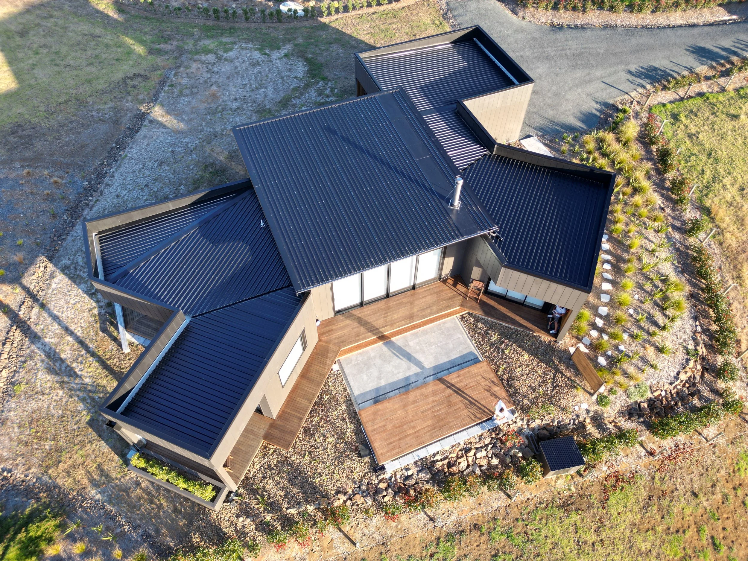 Aerial view of a modern, angular house with a dark metal roof surrounded by landscaped gardens and a gravel driveway.