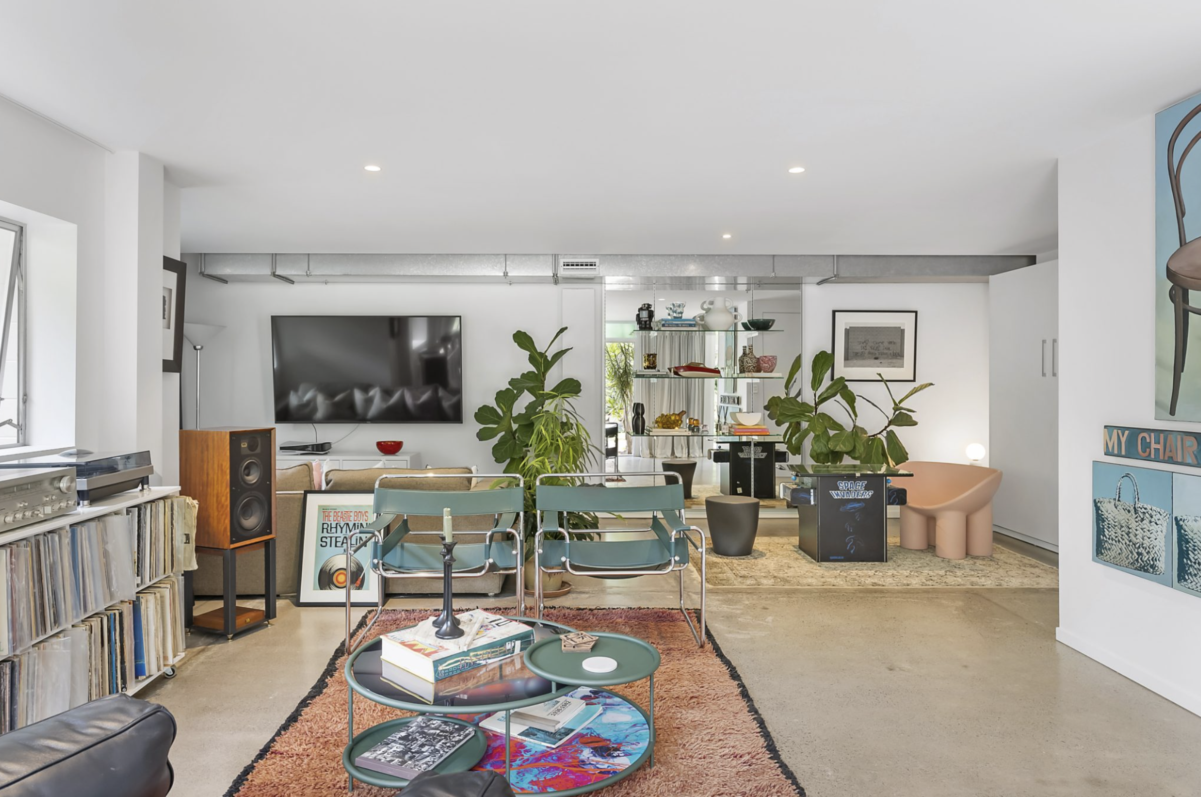 Modern living room with eclectic decor, featuring a flat-screen TV, vinyl record shelves, speakers, green chairs, round coffee tables, large potted plants, and artwork on the walls.