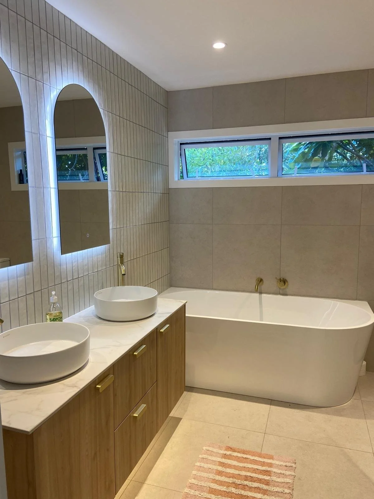 Modern bathroom with double sinks, large mirror, freestanding bathtub, wood cabinetry, and striped rug.
