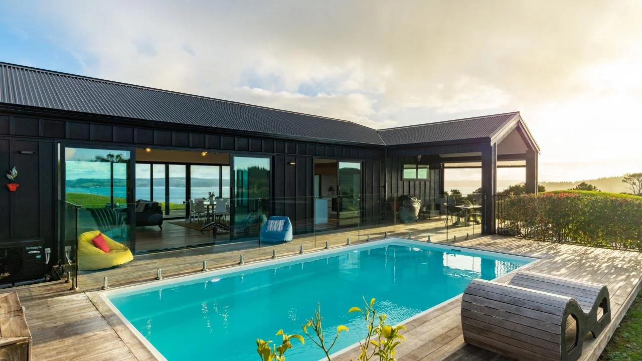 Modern house with glass, pool, outdoor seating, and ocean view at sunset.