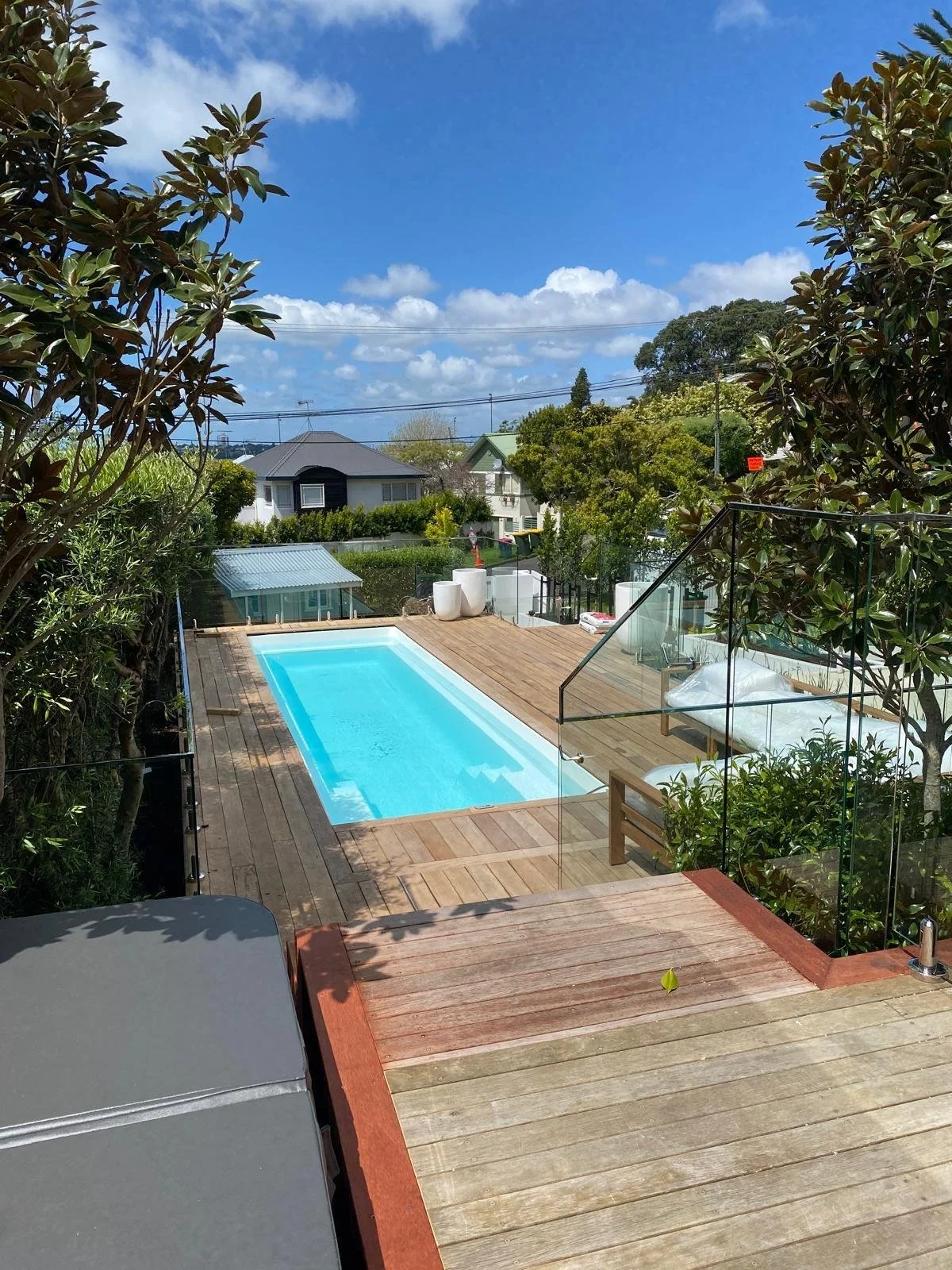 Backyard with a rectangular swimming pool, wooden deck, glass railing, and surrounded by trees and bushes, under a clear blue sky.