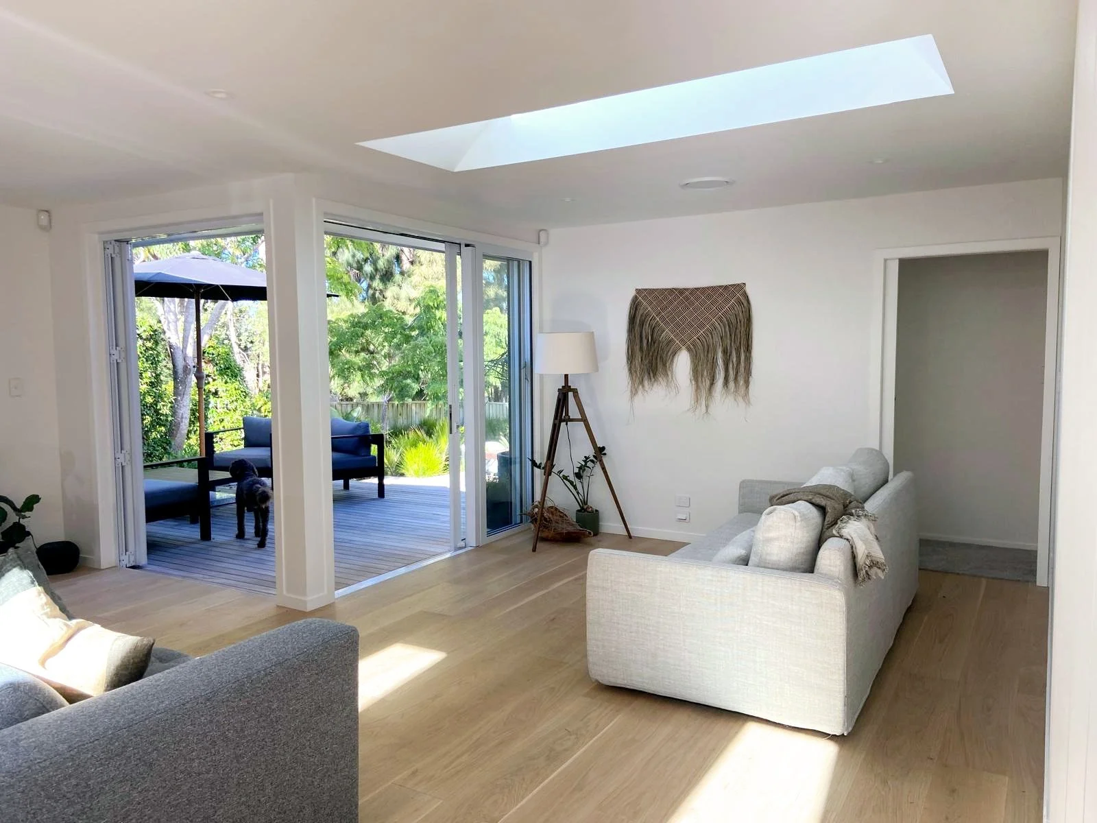 Living room with light gray sofas, wooden floor, and a skylight; sliding glass doors open to a deck with outdoor furniture and a dog; a wall hanging and a floor lamp are visible.