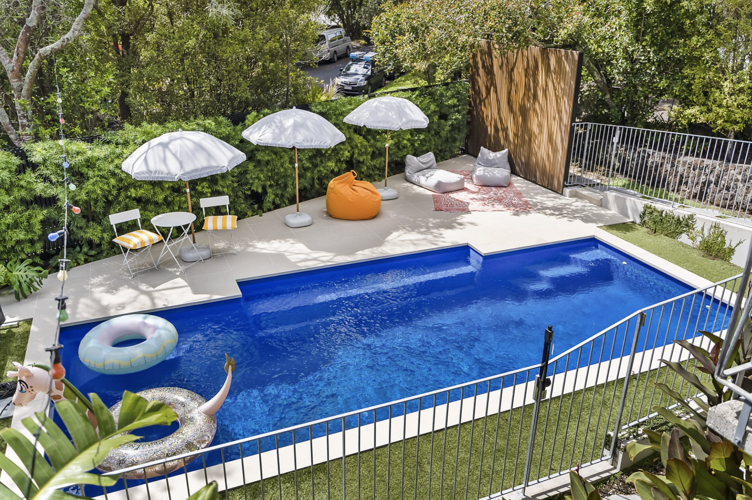 Backyard swimming pool with inflatable rings, surrounded by lounge chairs, umbrellas, a bean bag, plants, and a privacy fence.