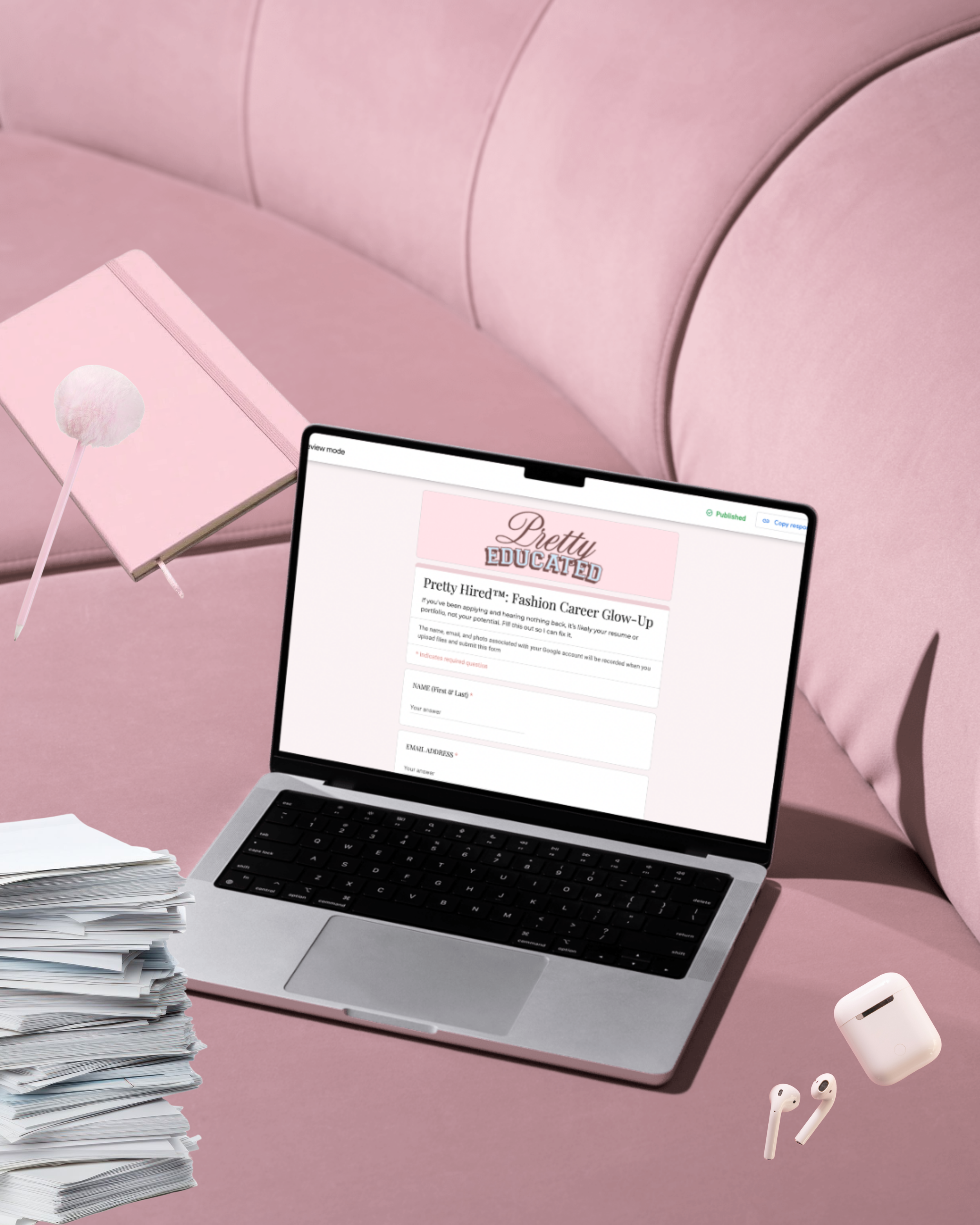 A workspace with a pink theme. It features a pile of papers on a desk, a pink notebook with a fluffy stick, a laptop displaying a website titled 'Pretty Hired,' and wireless earbuds, a charging case, and a pair of AirPods.