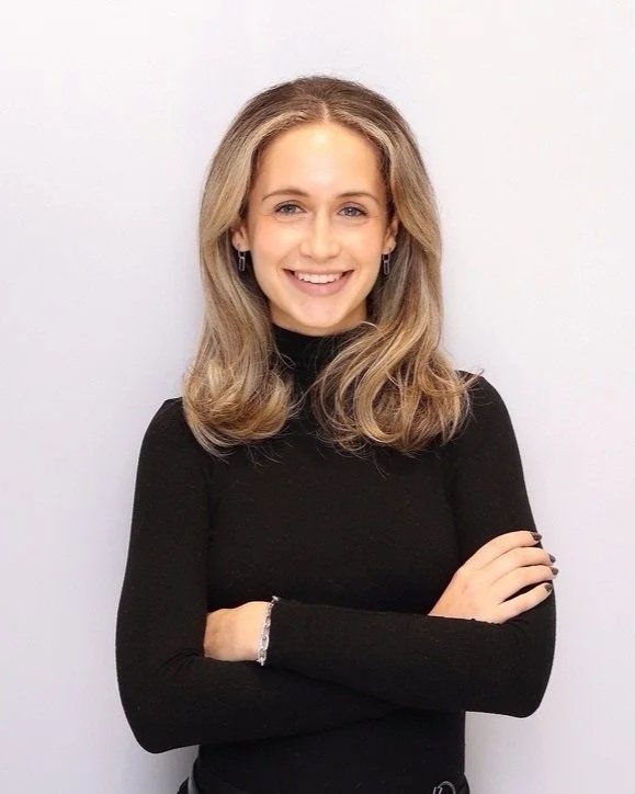 A woman with blonde hair and blue eyes wearing a black turtleneck sweater, smiling, with arms crossed, standing against a white background.