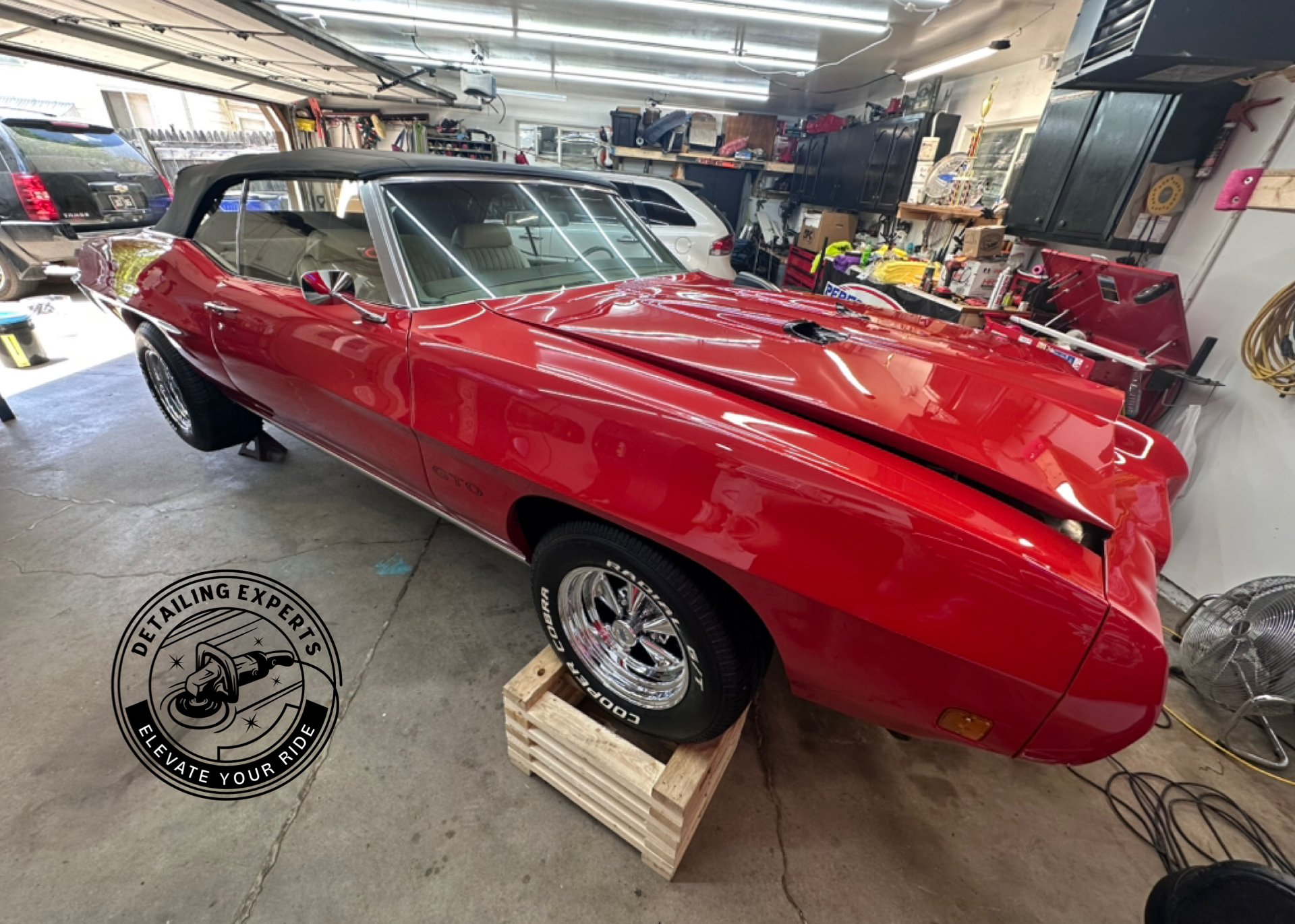 Red classic convertible car in a garage with a black soft top, raised on wooden blocks, surrounded by tools and equipment.