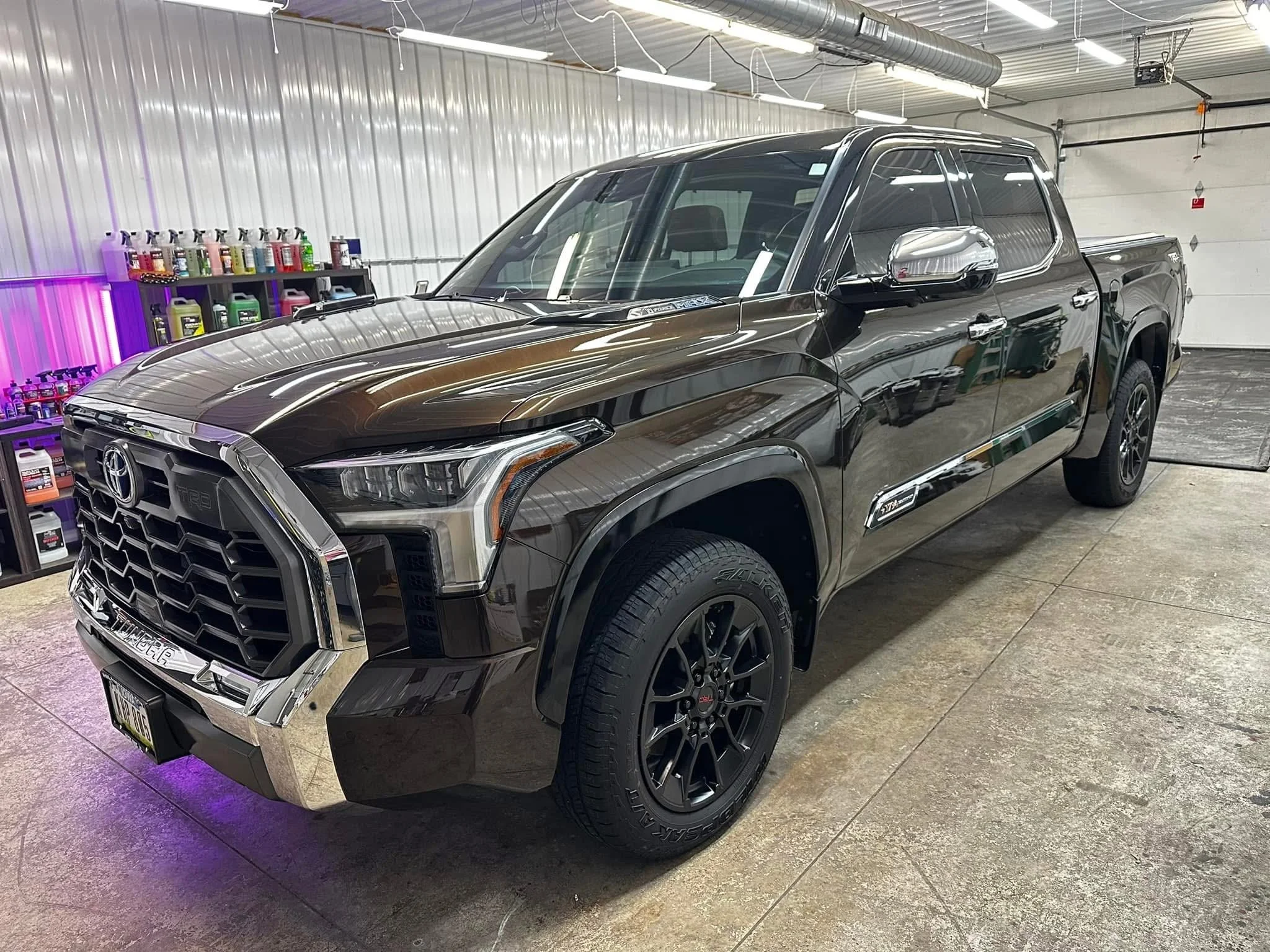 Black Toyota Tundra pickup truck parked inside a garage with a metal ceiling, shelving with various bottles, and tools visible in the background.