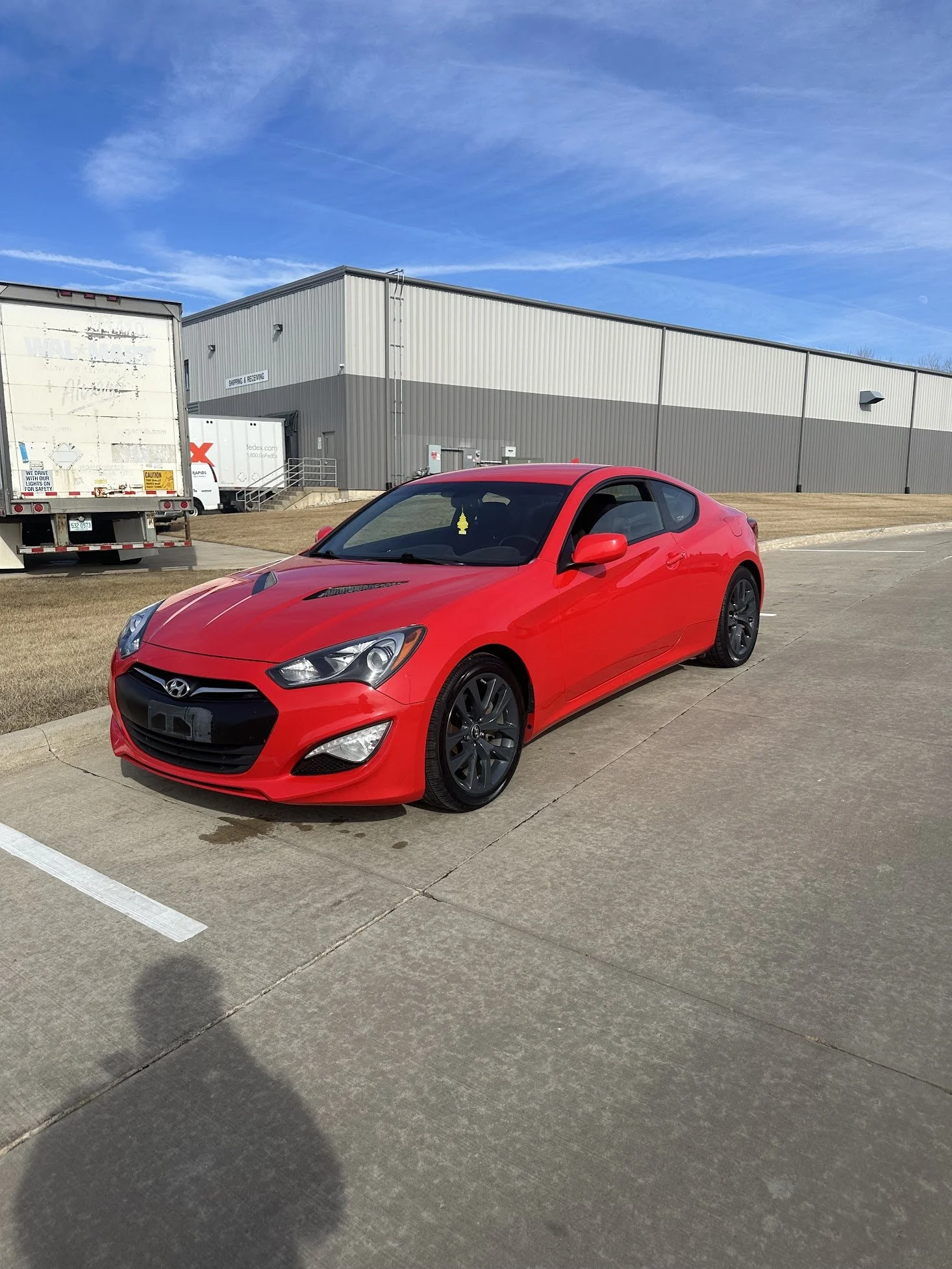 Red Hyundai coupe parked on a concrete surface in an industrial area with a large warehouse and trucks in the background under a blue sky.