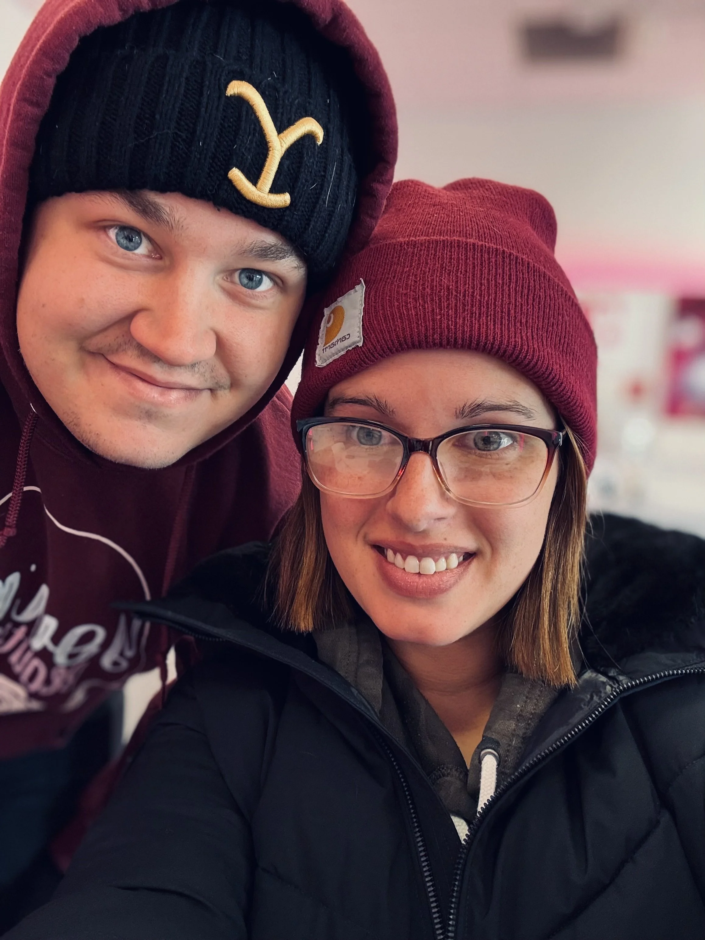 A man and woman taking a selfie together, both wearing beanies. The man has a black beanie with a yellow logo and is dressed in a maroon hoodie. The woman has a red beanie and wears glasses and a black jacket. They are indoors with a blurry background.